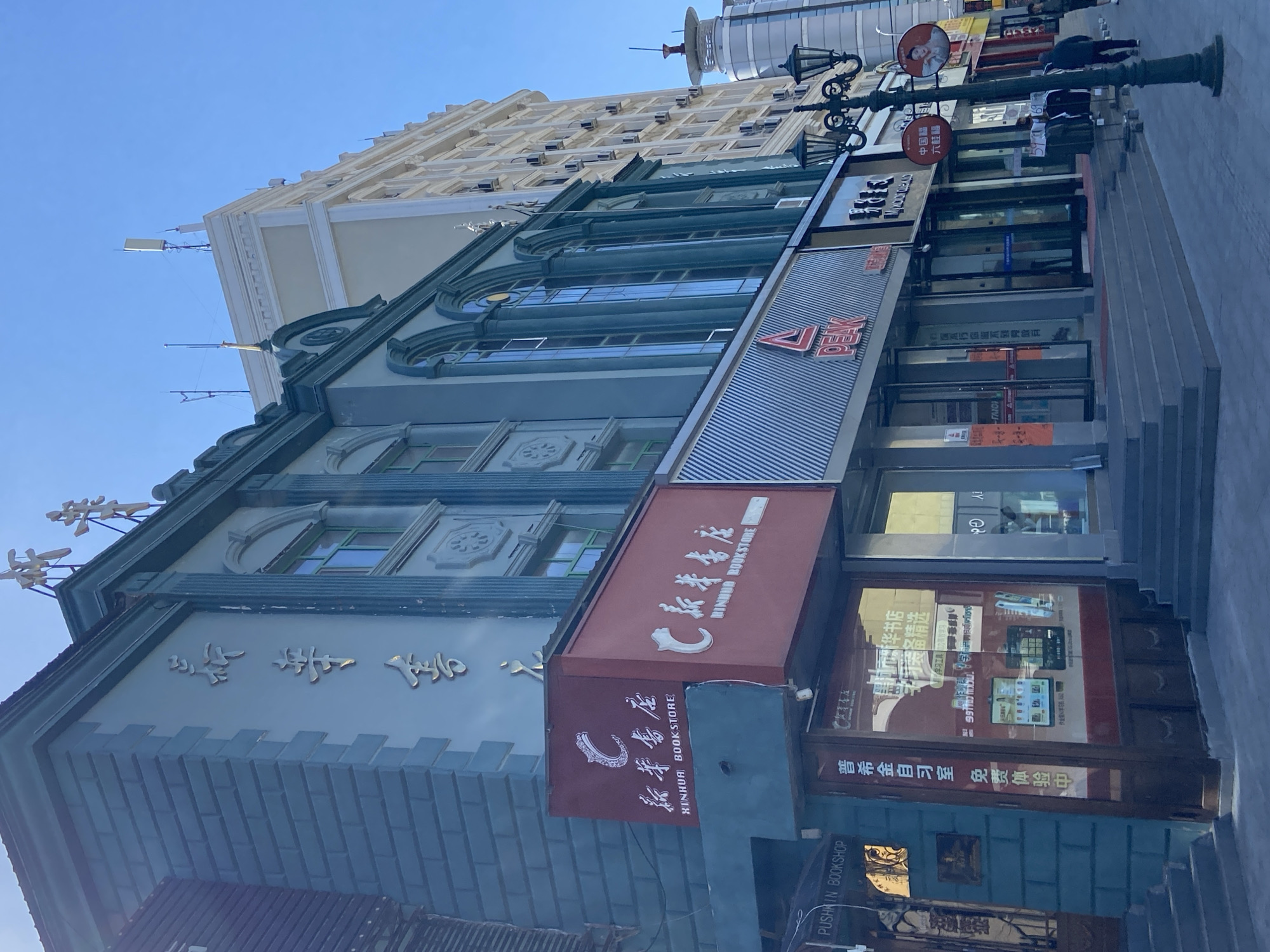 The image shows a street scene in Xing'an Jiedao, China, on a bright, clear day. The foreground features a row of shops with modern storefronts and traditional architectural elements. A prominent "XINHUA BOOKSTORE" sign in red and white is visible on the left, with Chinese characters above it. Adjacent to it, a shop with a blue and grey brick facade displays a sign that reads "PUSHCIN BOOKSHOP" in English. Further to the right, a storefront with a "PEAK" logo and "CHINA" signage is visible, along with other businesses. A black, ornate lamppost stands on the sidewalk, casting shadows. Several people are walking on the sidewalk in the mid-ground, dressed in casual clothing. In the background, taller buildings with numerous windows and air conditioning units are visible, indicating an urban environment. The sky is a clear, bright blue.