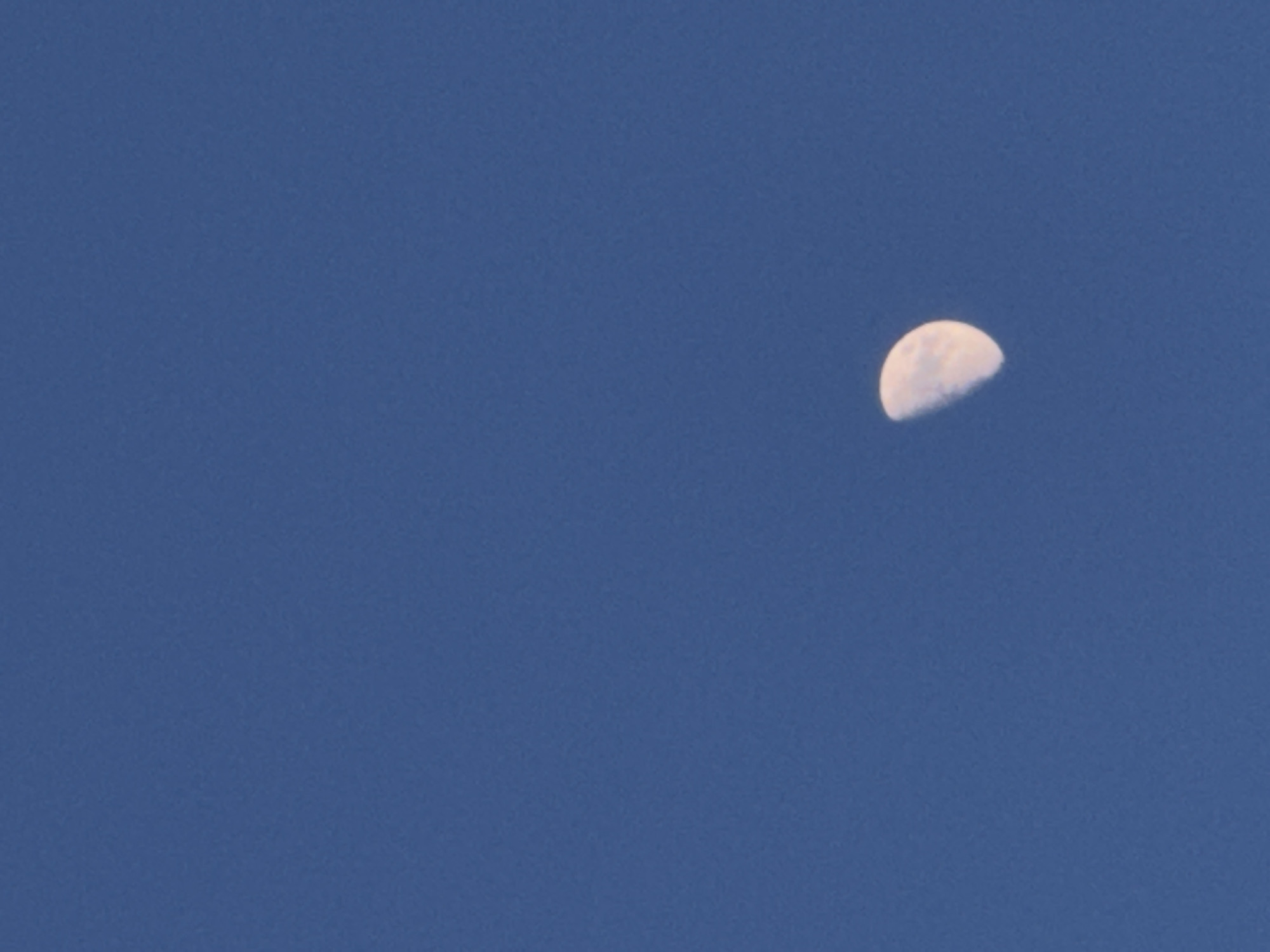 A partially illuminated moon is visible against a clear, deep blue sky. The moon appears to be in its waxing or waning gibbous phase, with roughly half of its disc illuminated. The sky is devoid of clouds and any other celestial bodies. The image is a close-up of the moon and sky, with no surrounding landscape or city elements visible.