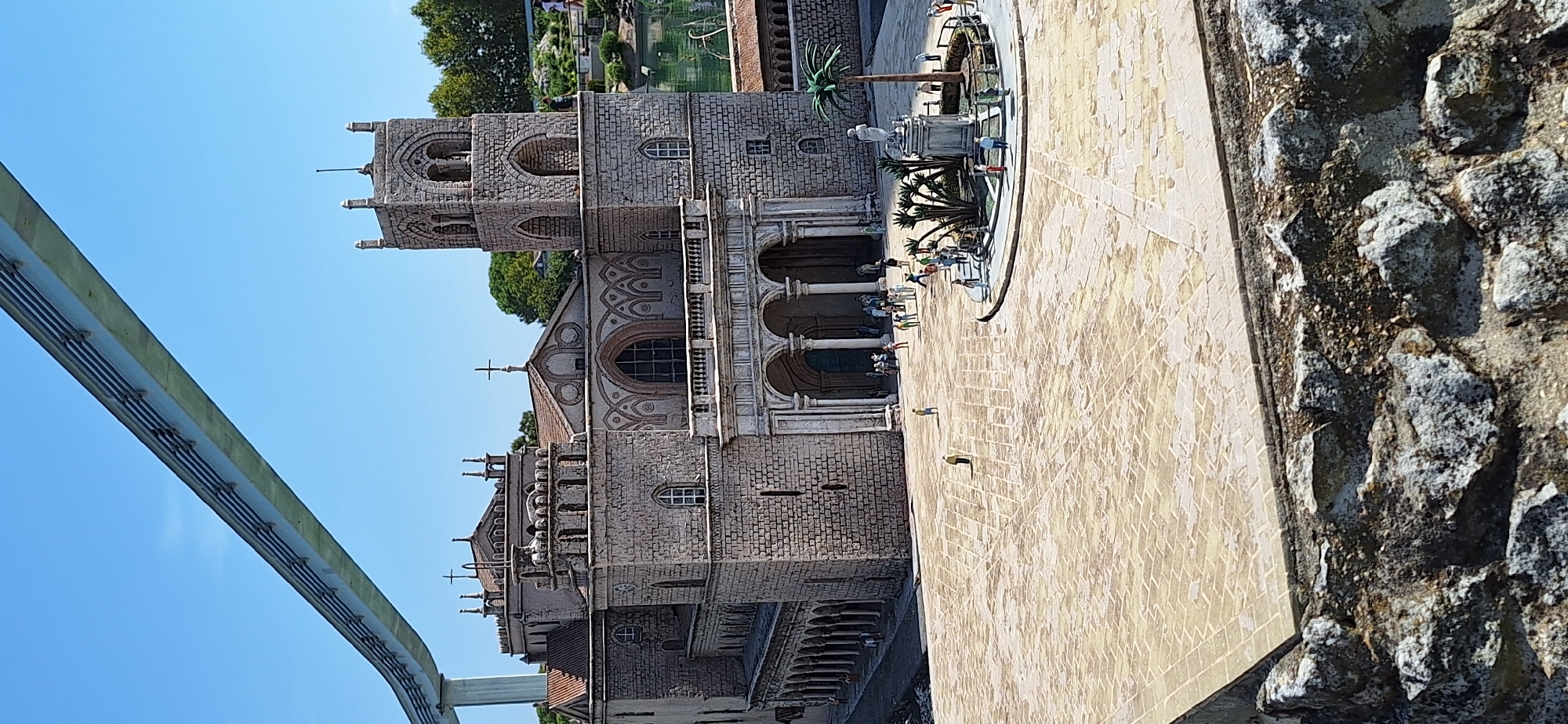 This is a detailed photograph of a miniature model of the Cathedral of Rimini, located in the Fiabilandia amusement park in Rimini, Italy. The model faithfully replicates the cathedral's stone facade, arched windows, bell tower, and decorative elements. In the foreground, a paved plaza surrounds the model, with several miniature figures of people scattered throughout, suggesting a bustling scene. A small fountain with palm trees and a statue is visible in the center of the plaza. The backdrop includes a clear blue sky and lush green trees, creating a vibrant and realistic setting for the miniature city. A monorail track is visible in the upper part of the image, indicating the presence of other attractions within the amusement park. The overall impression is one of meticulous craftsmanship and a playful representation of the historic landmark.