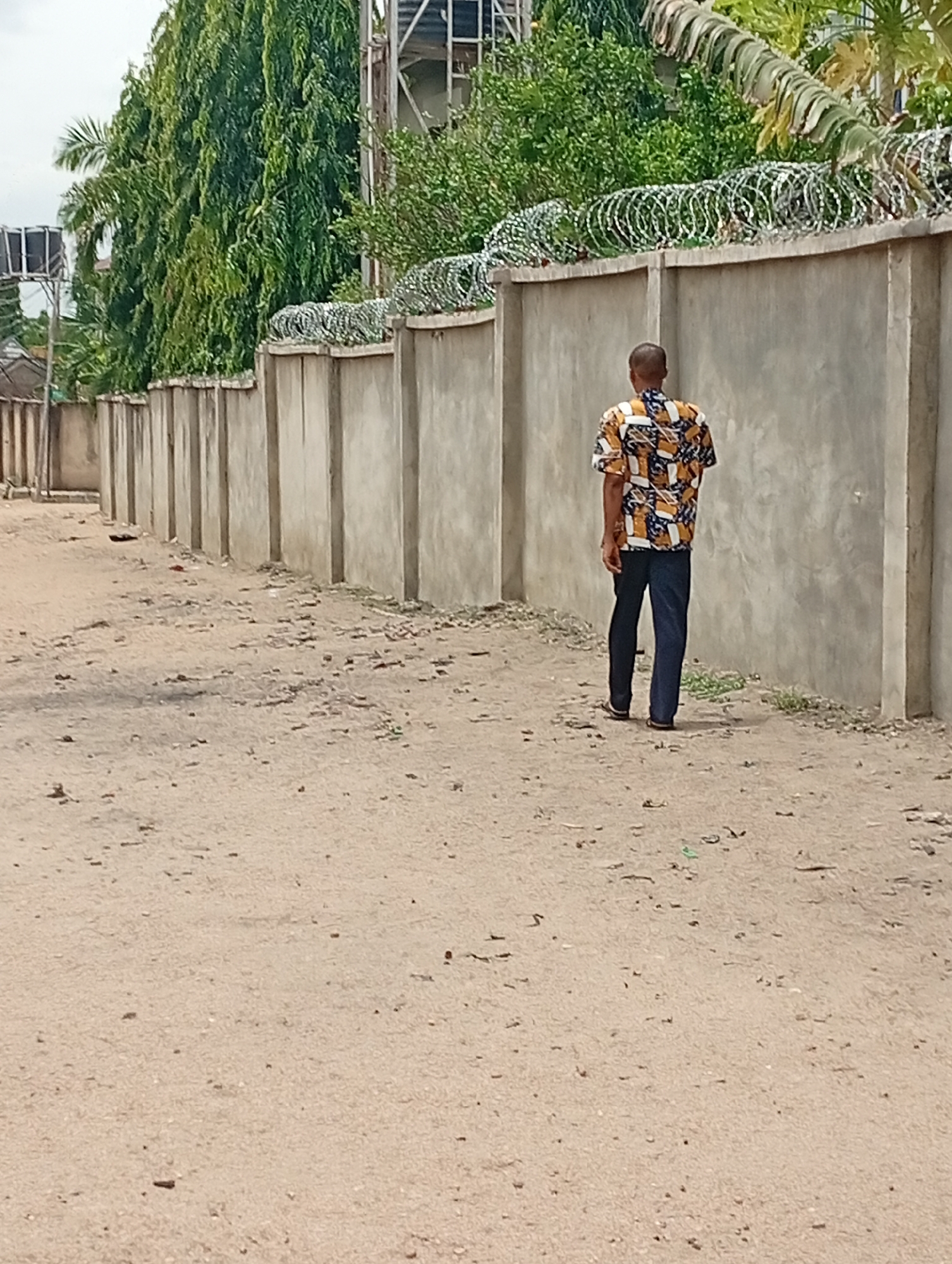 The image shows a man walking away from the camera on a dirt road in Adankolo, Nigeria. He wears a patterned short-sleeved shirt and dark pants. A tall concrete wall topped with barbed wire runs along the right side of the road, and trees are visible behind the wall. The ground is sandy with debris scattered across it.