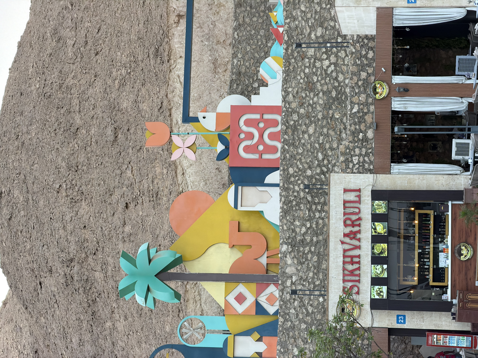 This image shows a street scene in Sharm El Sheikh, Egypt, featuring a hillside decorated with colorful abstract art and two restaurants. The art installation includes stylized palm trees, camels, birds, flowers, and geometric shapes, set against a rocky mountainside. Below the art, two restaurants are visible. The left restaurant, labeled "Sikhvaruli," appears to be a Georgian food restaurant and cafe, with a display of food items and shelves stocked with bottles. The right restaurant, also possibly a Georgian food establishment, has white curtains and outdoor seating. A Coca-Cola refrigerator is visible to the left of the Sikhvaruli restaurant. The environment suggests an outdoor dining area with a backdrop of natural rock formations.
