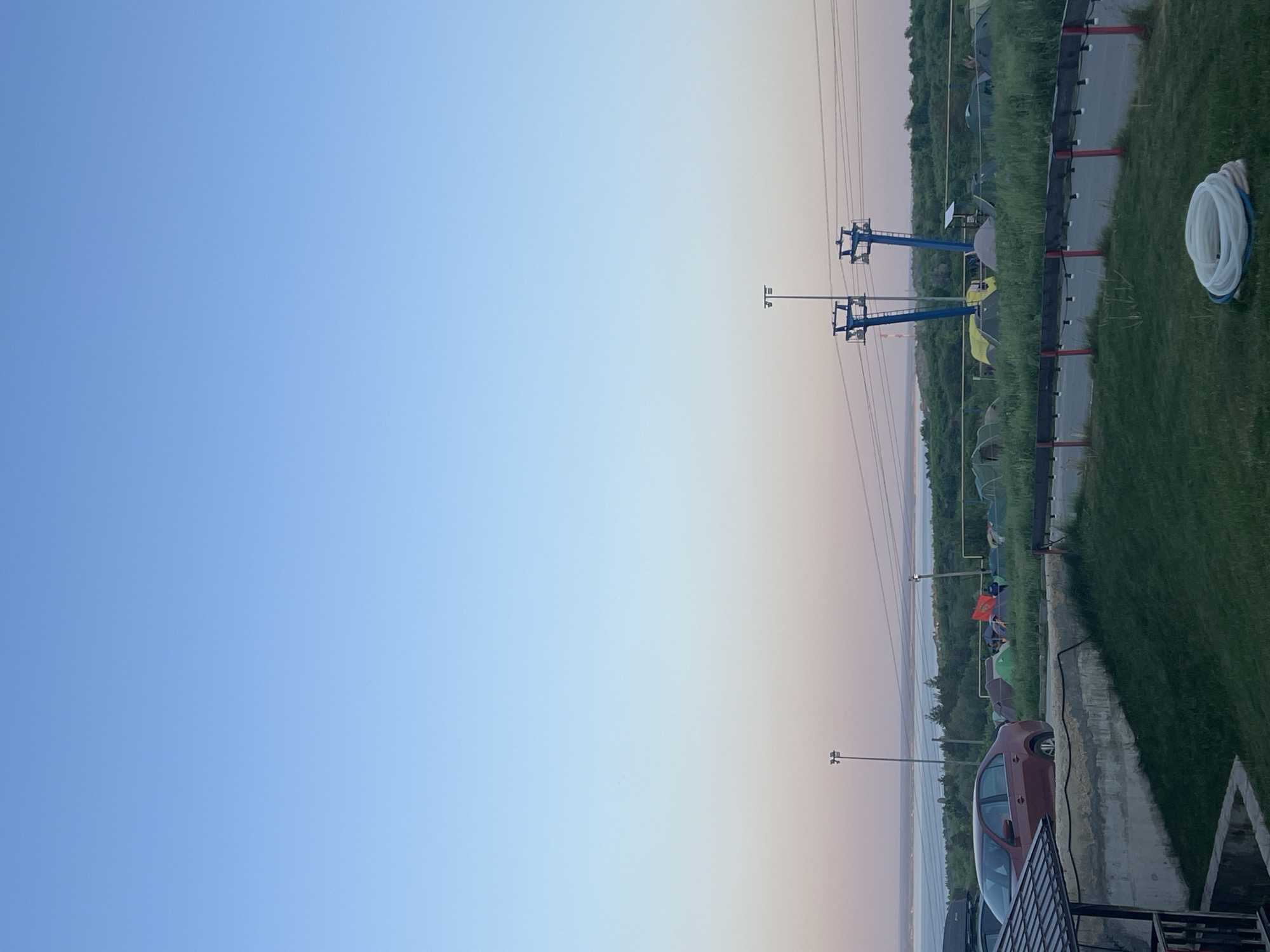 The image shows an outdoor scene in Khvalynsk, Russia. The sky is blue at the top, transitioning to light pink and white near the horizon. There are power lines extending from the top center of the frame to the bottom. The right side of the image features green grass, and a sidewalk with a low barrier, near tents, a car, and a coiled white hose. The scene includes chairlift towers.