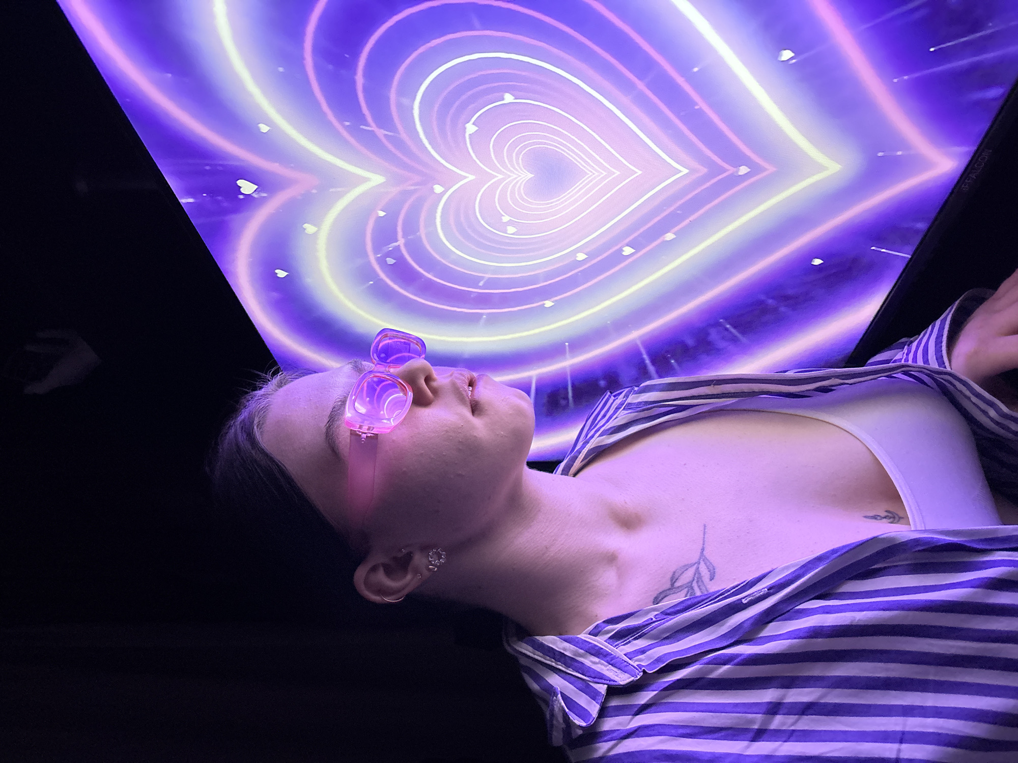 A person, likely a young adult female, is shown lying on their back in a dimly lit or dark room, looking upwards. They have short hair and are wearing distinctive pink-tinted glasses, which are pushed up onto their forehead, resting above their closed eyes. The individual is dressed in a striped button-up shirt (purple/blue and white) worn over a white tank top, with subtle leaf-patterned tattoos visible on their upper chest/shoulder area.
Above them, a vibrant and immersive light installation is displayed, possibly on a ceiling or large screen. This installation features a tunnel-like design created by numerous concentric heart shapes glowing in shades of purple, pink, and yellow neon lights. Smaller white heart-shaped particles appear to be floating within this light tunnel. The overall effect is dreamlike and ethereal, with the dark surroundings emphasizing the glowing heart pattern.
The person's posture and closed eyes suggest a moment of relaxation, contemplation, or immersion in the visual experience. In the upper right corner of the light display, faint white text reads "@iFFALCON". The scene takes place in Perm, Russia, likely within an art exhibit or a themed entertainment venue.