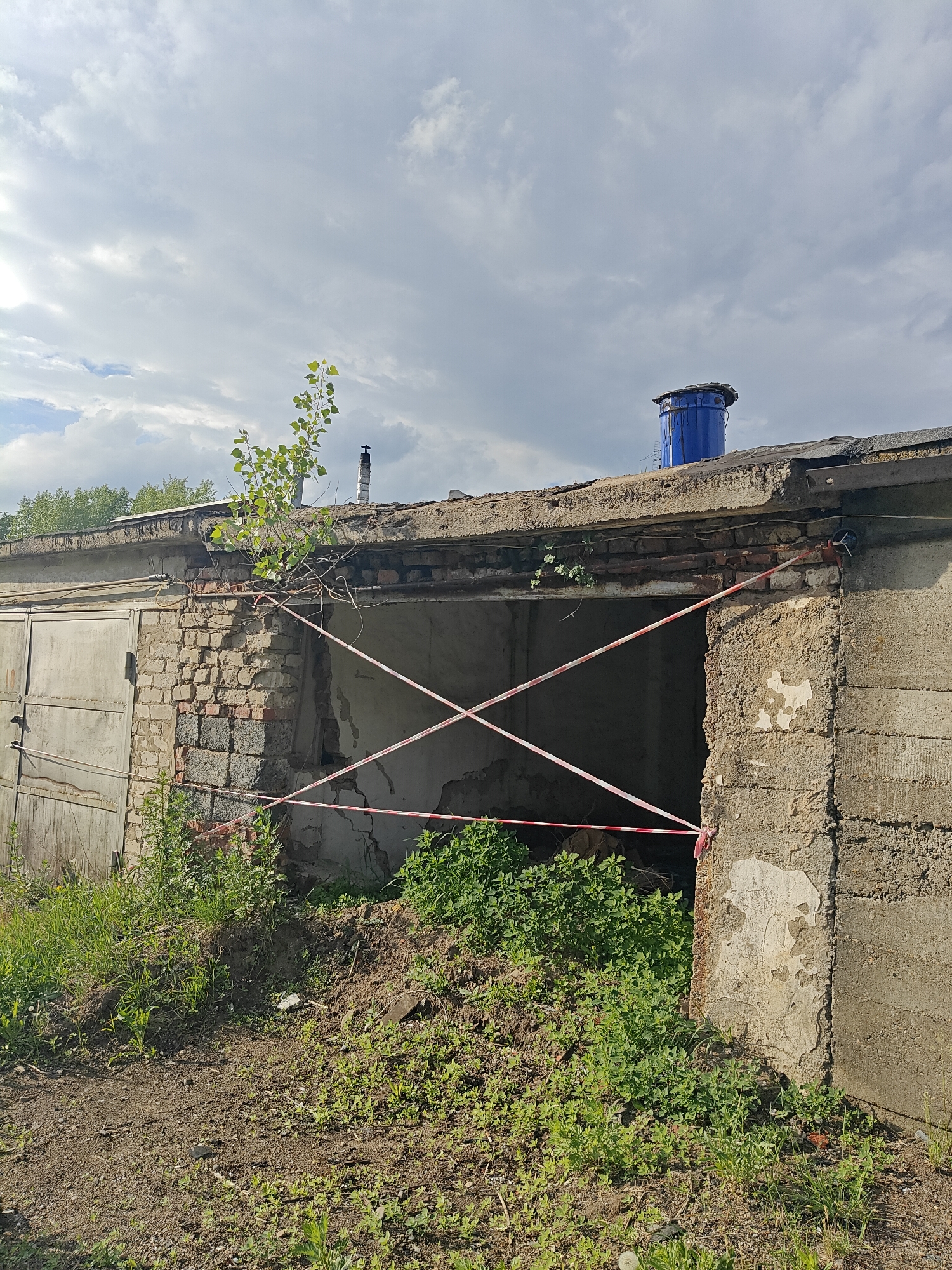 The image depicts a dilapidated, single-story brick and concrete garage structure in an apparent state of disrepair. The location is in Chaikovsky, Russia. The garage has a metal sliding door on the left and an open doorway on the right, demarcated with red and white caution tape. The brickwork of the wall adjacent to the open doorway is crumbling, and vegetation is growing in front of and on top of the structure. A small tree is growing from the roof near the edge. A blue container is visible on the roof. The surrounding area appears overgrown with weeds and grass. The sky is overcast.