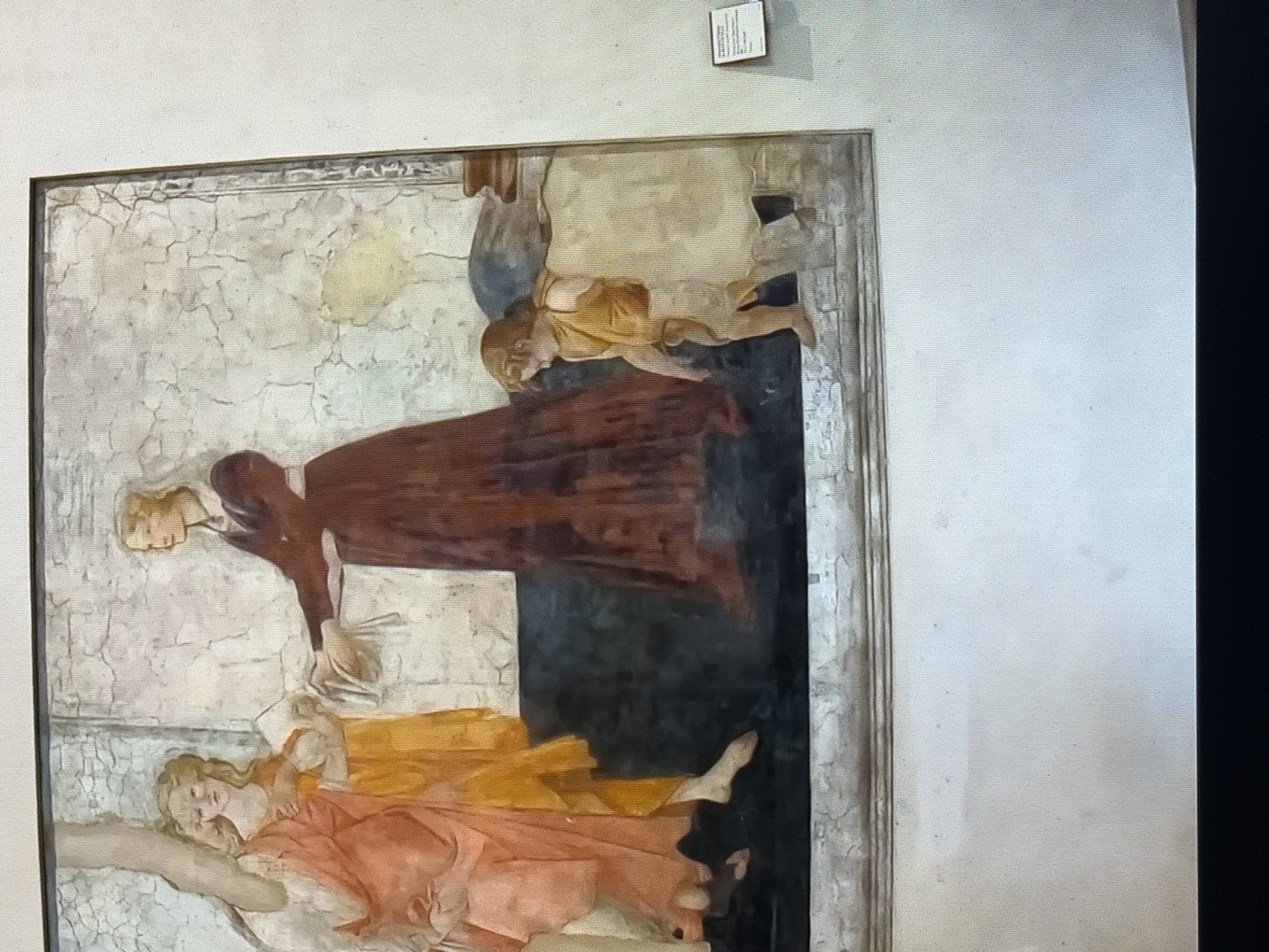 The image is a photograph of a fresco painting. The fresco depicts a scene with at least four figures. On the left, two female figures are shown in flowing orange and yellow garments. They appear to be interacting with a third female figure dressed in a dark red, floor-length gown and a white head covering. This third figure is extending her hands towards the first two figures, possibly in exchange for something. To the right of the main group, a young child is depicted walking away from the viewer, wearing a simple yellow tunic. The background of the fresco is a cracked, pale wall with hints of architectural elements. The fresco is framed by a decorative border at the bottom. A small, white informational plaque is visible on the wall to the right of the fresco. The overall lighting suggests an indoor setting, possibly a museum or gallery. The city and specific location are unknown.
