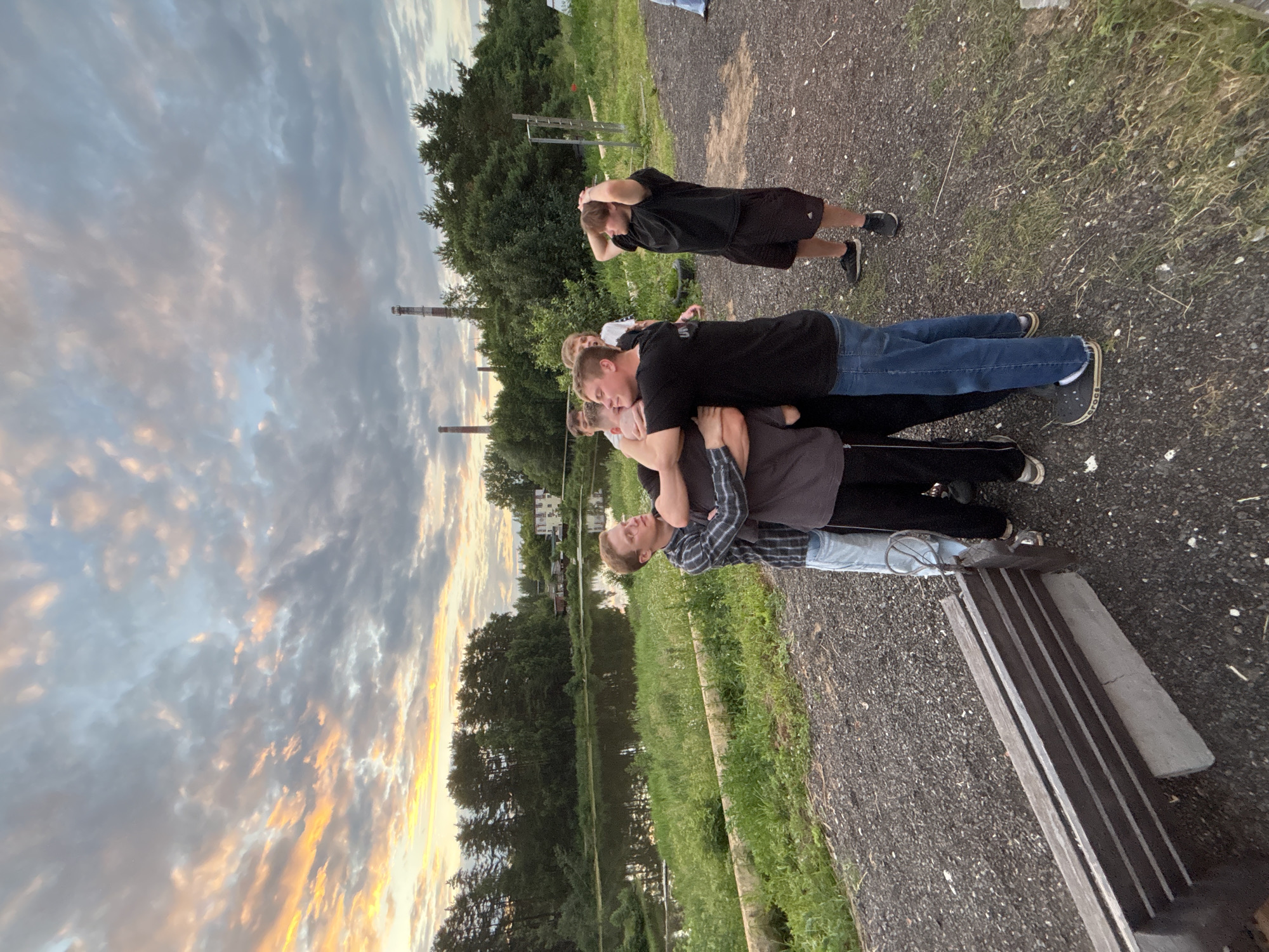 The image shows a group of young men in an outdoor setting with a lake and trees in the background, under a cloudy sky. In the foreground, four young men are huddled together, appearing to be hugging or engaging in some sort of playful roughhousing. One man on the right, wearing a black t-shirt and black shorts, stands slightly apart with his hands on his head, looking away. Another man, wearing a blue and grey plaid shirt and jeans, is on the left side of the huddle. The ground is covered in gravel, with some grass and vegetation visible. In the distance, industrial chimneys are visible above the trees, and a building can be seen across the lake. The overall impression is of a group of friends enjoying a late afternoon or early evening gathering.
Based on the presence of industrial chimneys typical of a thermal power plant and the specific architecture of these chimneys, it is highly likely that this scene is located near the Chaikovsky Thermal Power Plant in Chaikovsky, Russia. The landscape, with a lake and surrounding trees, is consistent with the natural environment around the industrial site in Chaikovsky.