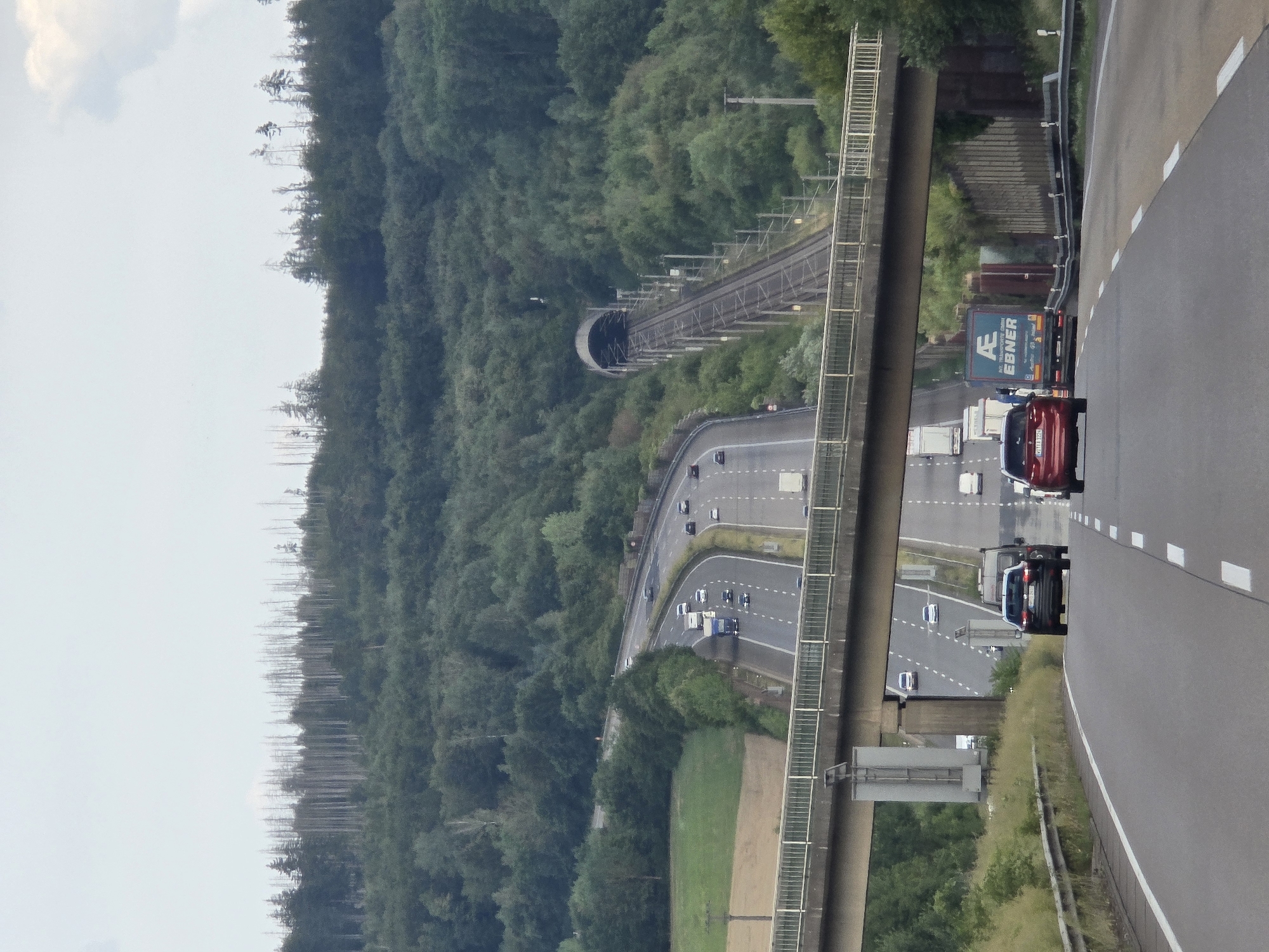 The image shows an aerial view of a highway and a train tunnel through a forested hillside, likely in Deesen, Germany. The highway is paved with asphalt and marked with white lane lines. Several vehicles are on the highway, including cars, trucks, and vans. One truck is blue and has the text "AE EBNER" printed on its side, along with a logo. Another truck is silver, and a red car is visible. The train tunnel is curved and appears to be made of concrete. Train tracks lead into and out of the tunnel. The surrounding area is covered in dense green trees, with some bare trees visible on the upper part of the hill. The sky is overcast with some clouds. The overall scene depicts a transportation infrastructure integrated into a natural landscape. There are no people clearly visible. The time of day appears to be daytime, and the weather seems to be cloudy.