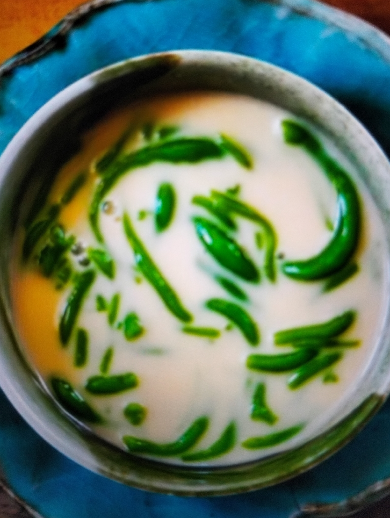 A close-up, overhead shot showcases a bowl of a dessert, likely Cendol, a popular Indonesian sweet treat. The dessert consists of green, jelly-like strands suspended in a creamy, white liquid, possibly coconut milk or sweetened milk. A yellowish, translucent liquid is visible at the bottom of the bowl, which could be palm sugar syrup. The bowl itself appears to be a light gray or white ceramic, and it rests on a decorative, turquoise-colored plate with a rough texture. The background is blurred, suggesting an indoor setting with warm lighting. There are no people or specific identifiable landmarks of Payagaboh, Indonesia present in this image; the focus is solely on the dessert.