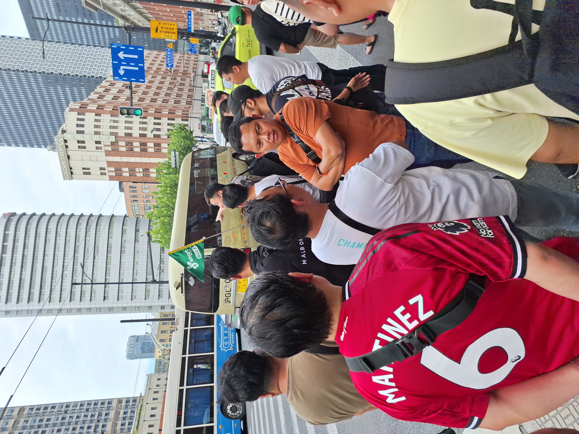 A crowd of men are gathered on a street corner, with a yellow bus and a blue bus visible in the background. The men are of various ethnicities and are dressed in casual clothing. Some are wearing t-shirts, while others are wearing collared shirts. One man in the foreground is wearing a red jersey with the number 6 and the name "MARTINEZ" on the back. He also has a black strap across his chest. Another man, wearing an orange t-shirt, has his arms crossed. A green flag with a "Smart Mobile" logo is visible on the yellow bus. Traffic lights and street signs are present, indicating an urban environment. The buildings in the background are tall and modern, some with distinct architectural features, including a building with "GOLDEN TULIP" signage. The overall scene appears to be a public street in a city, possibly in China, given the language on the street signs.