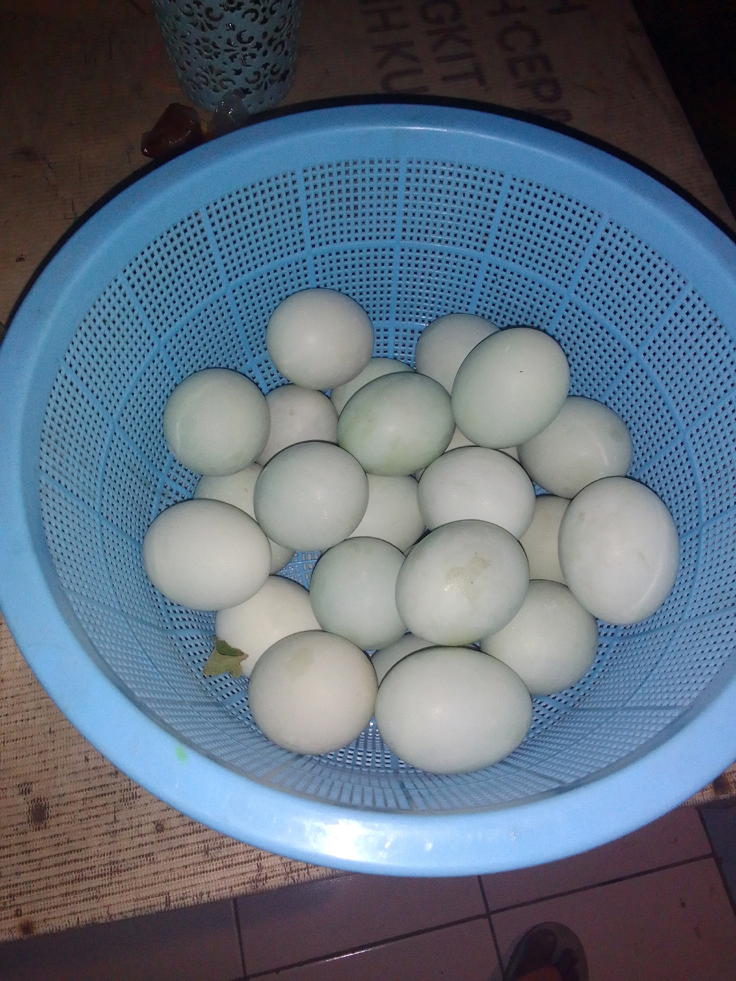 A blue plastic basket containing approximately 20 white eggs is positioned on a wooden surface. The basket is perforated, and the eggs are densely packed within it. The background is dark and out of focus, with some text visible on a flat surface behind the basket, though it is illegible. The overall scene appears to be an indoor setting.