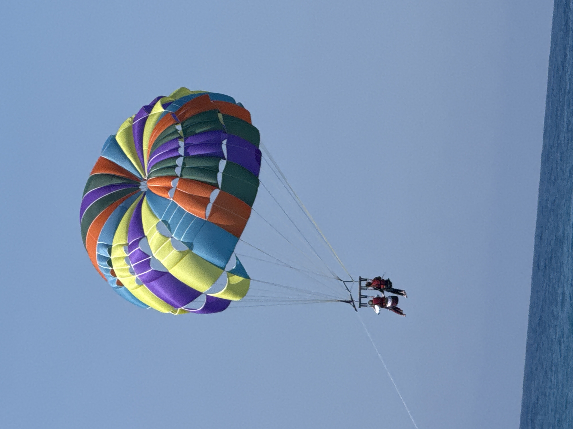 An eye-level, medium shot captures a colorful parasail with two individuals riding beneath it against a clear blue sky. The parasail is a vibrant mix of purple, yellow, green, and orange panels. Below the parasail, two people are seated in a harness, appearing to be enjoying a parasailing experience. The harness is suspended by multiple white lines from the parasail. In the lower portion of the image, the shimmering blue surface of the ocean meets the sky at the horizon, with a faint line of the Turkish coastline visible at the very bottom. The scene depicts a recreational activity common in coastal tourist destinations.