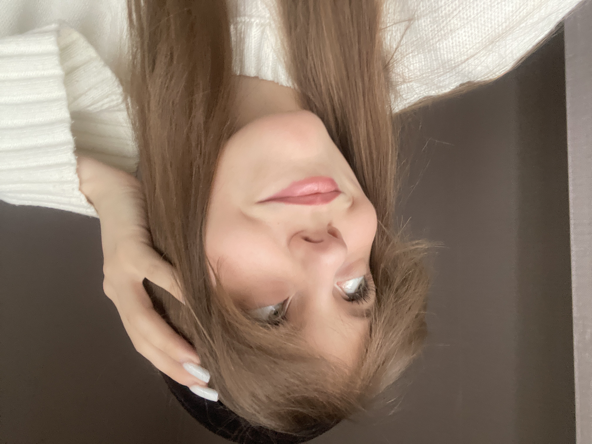 A female person is depicted from a low angle. Her face is visible, with light skin, brown hair, and light-colored eyes. She's wearing a white sweater. Her hand is visible, with white painted nails. The background appears to be a dark gray or brown wall. There is no indication of the location being the city of Ufa, Russia.