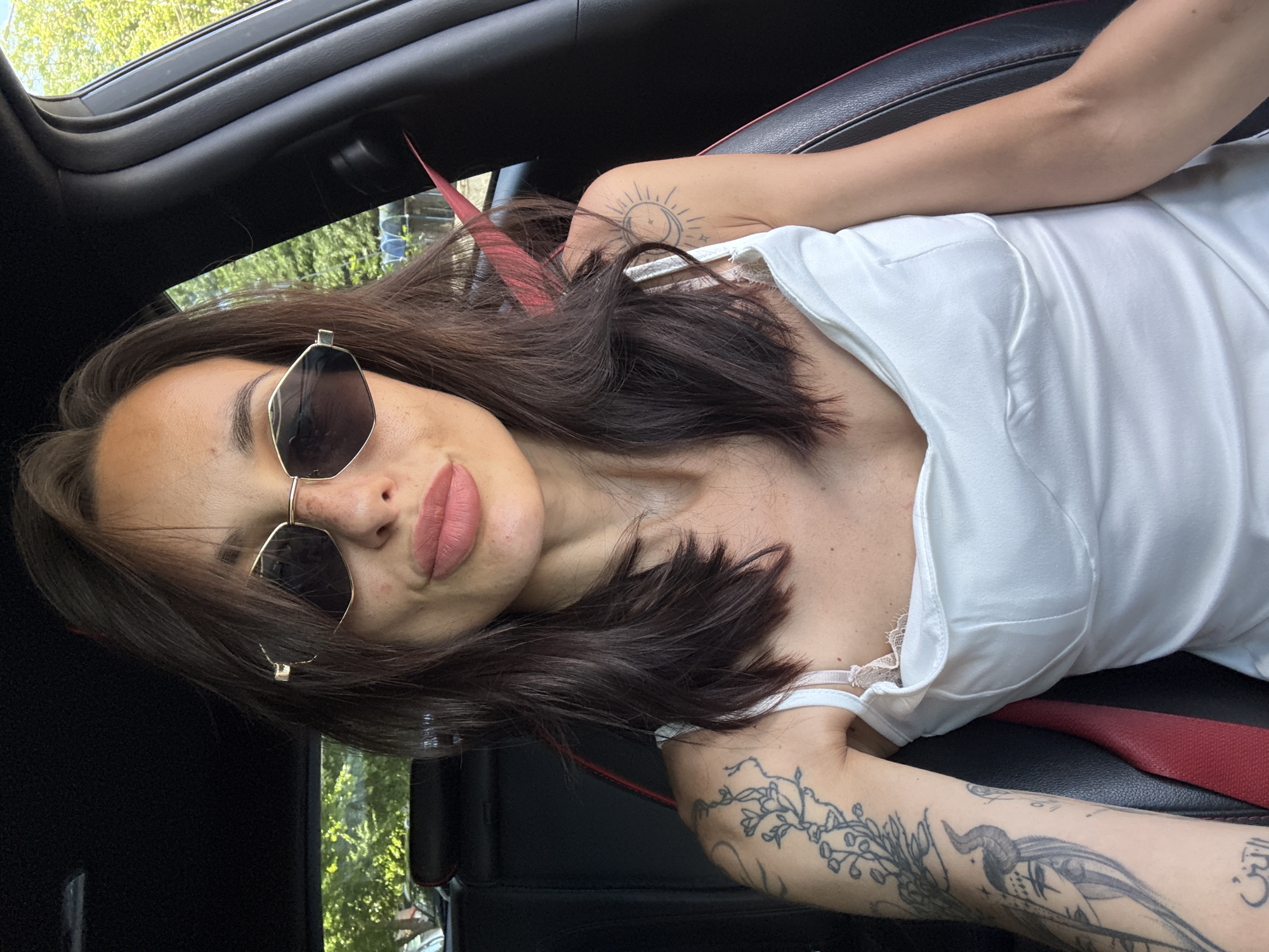 A woman is sitting in the driver's seat of a car, looking directly at the camera. She has dark brown hair, parted in the middle and falling to her shoulders. She is wearing sunglasses with hexagonal frames and a white camisole. There are tattoos on her left arm and shoulder. The interior of the car is visible, including a black leather seat with red trim and a red seatbelt. The background outside the car is blurred, showing trees and foliage, suggesting an outdoor location, possibly in a residential area or park within Pleven, Bulgaria. The image is taken from a slightly low angle, with the woman's face filling most of the frame.
