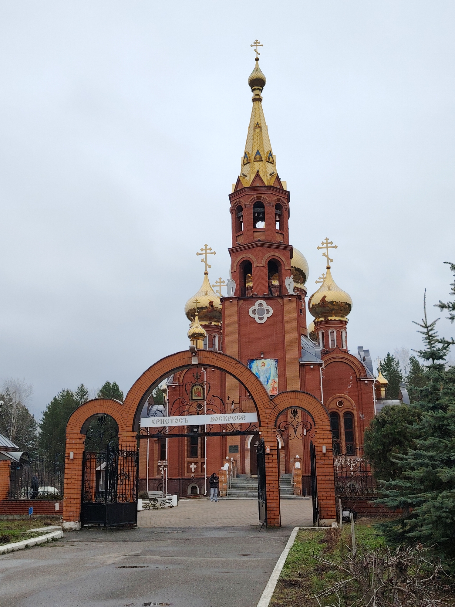 The image depicts the exterior of a red-brick Orthodox church in Chaikovsky, Russia, under an overcast sky. The church features multiple golden onion domes, each topped with an Orthodox cross, and a tall bell tower with a prominent golden spire.
An arched brick gateway with ornate black metal gates forms the entrance to the church grounds. Above this archway, a white banner displays the Cyrillic text "ХРИСТОСЪ ВОСКРЕСЕ!" (CHRIST IS RISEN!). The church facade includes a large fresco depicting the Virgin Mary with an infant and smaller icons. White angel statues are visible on ledges near the bell tower.
The paved ground leading up to the gate appears wet in areas, suggesting recent rain or damp conditions. Green coniferous trees are visible on both sides of the church and in the foreground, alongside some bare bushes and grass. Two individuals are present within the church compound: one stands near the church steps, and another further back to the left. A white bicycle is parked near the gate, and benches are placed near the church entrance. The scene portrays a serene, quiet daytime atmosphere at the religious site.