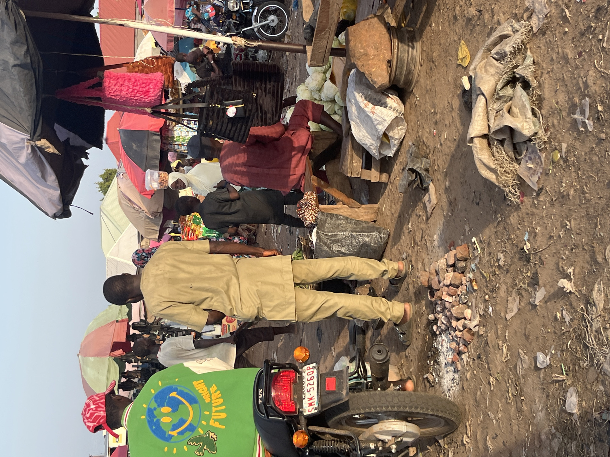 The image depicts an outdoor market scene. In the foreground, a motorcycle is parked on the left, with its rear wheel and license plate visible. The license plate reads "SMK-5260P". A pile of reddish-brown stones and debris is scattered on the dirt ground in front of the motorcycle. Several people are present, including a man in a green shirt with a smiley face and globe graphic, wearing a red and white patterned cap. Another man, seen from the back, wears a tan tunic and trousers, with sandals. A person in a burgundy outfit is seated on a wooden bench, with a pile of white produce behind them. Other individuals are visible in the background, some under umbrellas, and a variety of goods are displayed. The environment appears dusty and worn, with debris scattered on the ground, suggesting a busy, functional market setting.