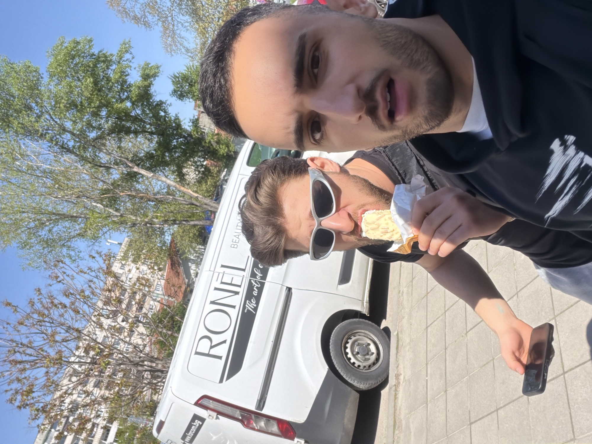 An outdoor selfie captures two men on a bright, sunny day, with the image rotated 90 degrees clockwise. The man in the foreground, with dark hair and a beard, looks directly at the camera with an open-mouthed expression, wearing a black hoodie over a white t-shirt. Behind him, another man with brown hair and a beard wears distinctive white-framed sunglasses and is in the process of eating a light-colored snack, possibly an ice cream bar. He holds a black smartphone in his right hand.
In the background, a white commercial van is prominently parked, partially obscuring a distant building and lush green trees. The van features the large, dark text "RONEL," with "BEAUTY" visible above it and "The art of e" (likely "The art of excellence") below it. A web address, "www.ircor.bg," is clearly visible on the lower rear of the van. The ground is paved with light-colored tiles or concrete, typical of a city street or sidewalk. The clear blue sky suggests good weather. The "ircor.bg" domain, combined with the context, indicates the location is in Bulgaria, specifically Plovdiv.