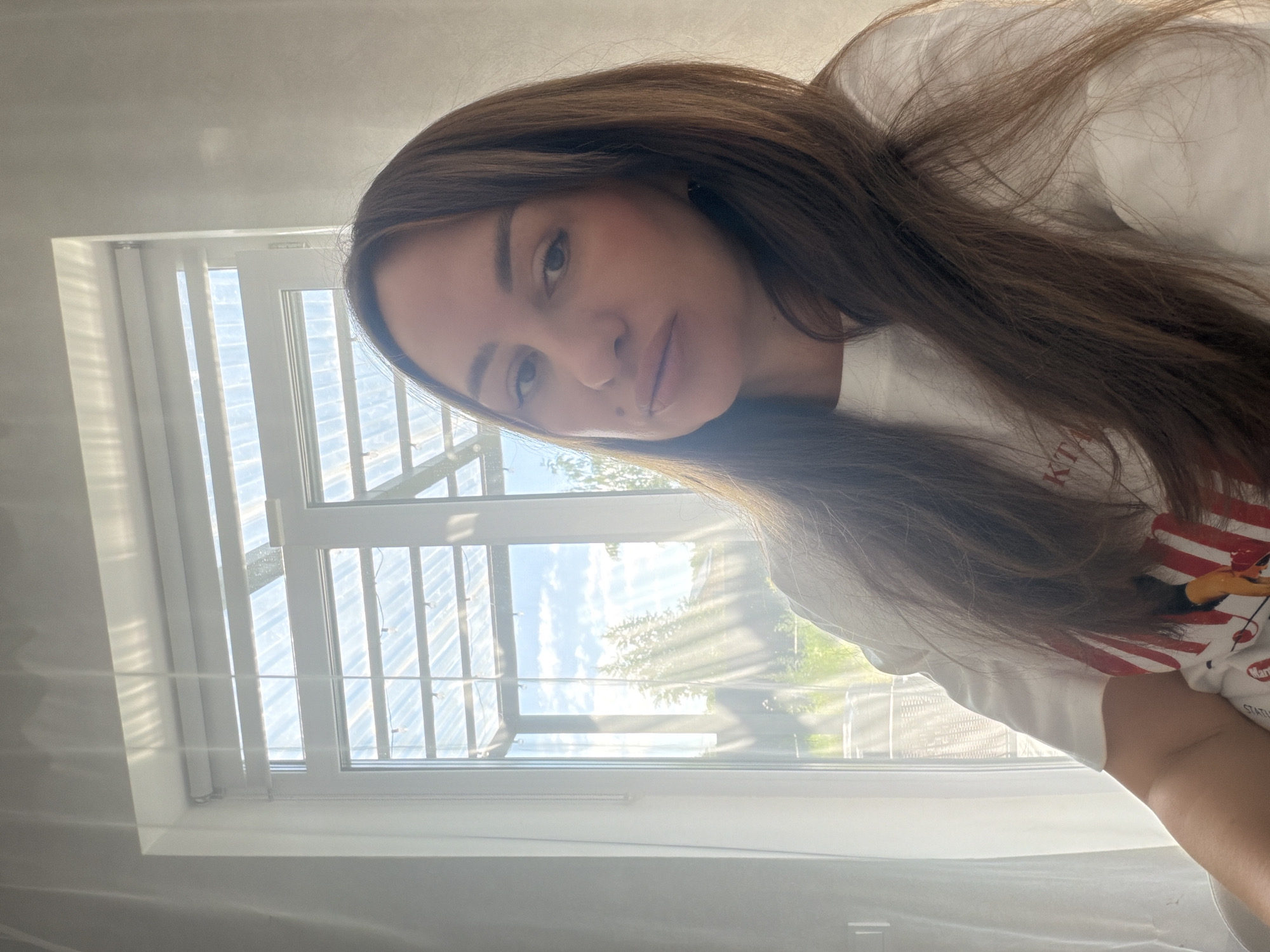 In Malinovka, Russia, a woman with long, brown hair sits indoors, facing the camera. She is wearing a white t-shirt. Behind her is a large window with blinds, which offers a view of a greenhouse-like structure with sunlight streaming through. The woman is positioned in the foreground, with the window and outdoor scenery in the background.
