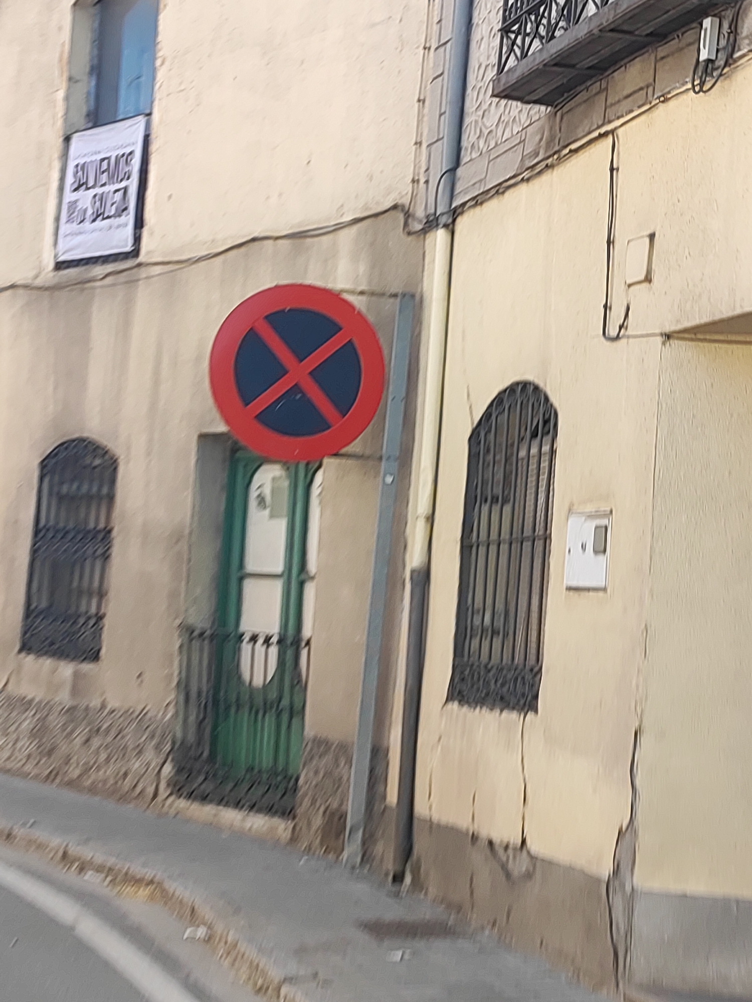 The image captures a street scene in Santa María La Real De Nieva, Spain. A weathered, light-colored building dominates the frame, featuring a dark green door with ornate metalwork, and two barred windows. A large, circular red sign with a blue cross inside, indicating a no parking zone, is prominently displayed on a pole to the right of the door. A white banner with black text, partially visible, hangs from a window on the upper left side of the building. The building's facade shows signs of age, with cracks visible in the plaster. A sidewalk and a street are visible in the foreground, with dry leaves scattered on the pavement. Another window with a black metal grate is visible on the left side of the building.
