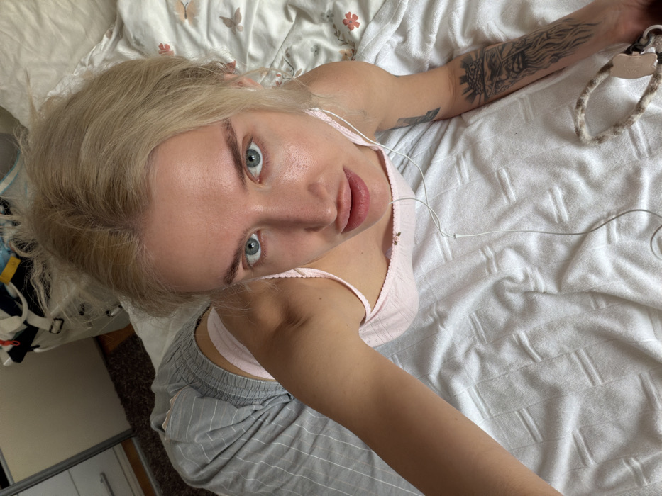 The image depicts a female subject, appearing to be an adult, in an indoor residential setting, likely a bedroom. The precise location within the city of Budva, Montenegro, is not discernible from the visual information provided. The subject has light blonde hair, blue eyes, and fair skin, and is positioned prone or kneeling on a bed, looking directly upward into the camera. She is wearing a light pink camisole and light grey striped pants. A tattoo of a bearded male figure's head, possibly crowned or wearing a headpiece, is visible on her left forearm. White wired earbuds are draped across her chest and abdomen. A braided or woven accessory is partially visible in the bottom right corner of the frame, near her right hand. The bed is covered with a white textured blanket, and a floral-patterned sheet or pillow is visible behind her head. In the background, a mirrored wardrobe door and miscellaneous cluttered items are visible. A dark-colored rug is present on the floor to the left.