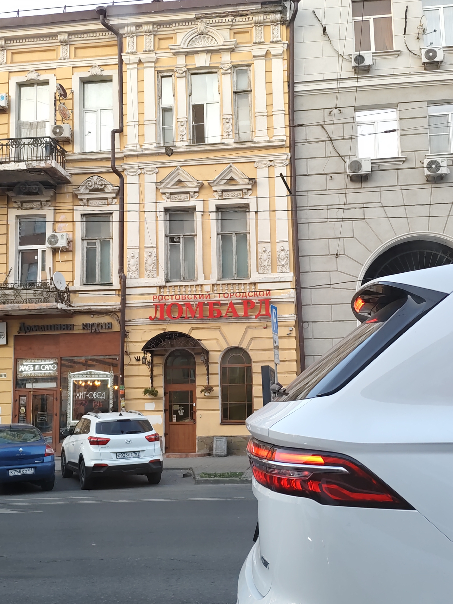 The image shows a street scene in Rostov-on-Don, Russia. In the foreground, the rear of a white car is visible, with its brake lights illuminated. The car is parked on the side of a paved road. Behind the car, there are several buildings. The building on the left is a yellow, multi-story structure with ornate architectural details, including columns and decorative molding around the windows. One of the windows has an air conditioning unit attached. There is a balcony on the second floor with a black metal railing. A red sign with Cyrillic lettering is prominently displayed on the yellow building, which appears to read "ЛОМБАРД" (LOMBARD) and "РОСТОВСКИЙ ГОРОДСКОЙ" (ROSTOV CITY). Below this sign, there are storefronts with signs like "ДОМАШНЯЯ КУХНЯ" (HOMEMADE KITCHEN) and "ХЛЕБ И САЛО" (BREAD AND SALO). To the right of the yellow building is a lighter colored, multi-story building with a more modern facade, also featuring multiple windows and air conditioning units. The sky is overcast, suggesting it might be a cloudy day.