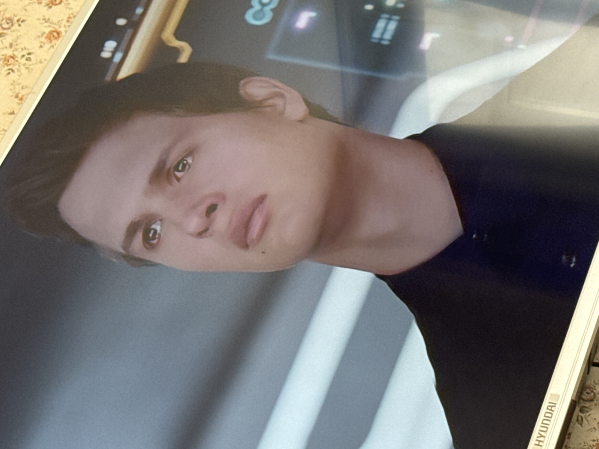 The image shows a framed portrait of actor Ansel Elgort, viewed from a tilted angle. The background within the frame is a blurred, futuristic-looking scene with lights and graphic elements. He is wearing a dark shirt. The bottom right corner of the frame reads "HYUNDAI" in capital letters. The surface the frame is resting on has a floral pattern. It is impossible to determine from the picture if the photo was taken in Mytishchi, Russia or whether it is in the city at all.