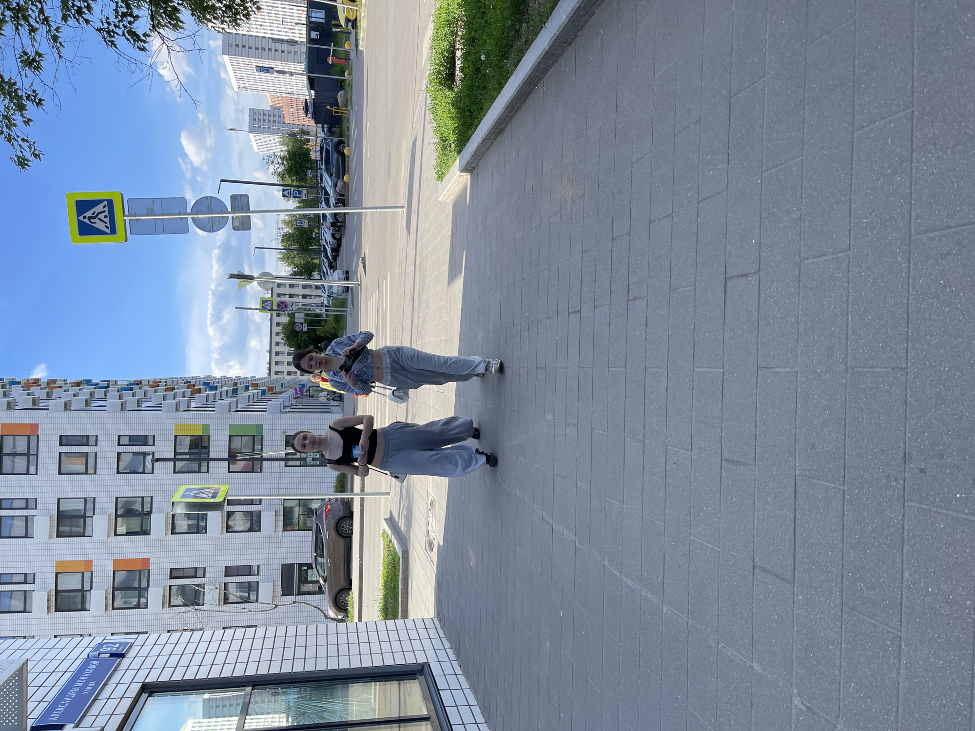 Two young women are standing on a paved sidewalk in an urban setting, likely Kommunarka, Russia, during bright daylight hours. The image is taken outdoors on a sunny day, with a clear blue sky dotted with some white clouds.
One woman, closer to the foreground, is wearing a black tank top, grey sweatpants, and dark shoes. She holds a small object, possibly a phone or water bottle, in her hands. The second woman, slightly behind and to the left, wears a light blue and white striped long-sleeved shirt, grey sweatpants, and light-colored sneakers. She carries a white handbag on her shoulder. Both women are standing and looking towards the camera, appearing to pose or be captured in a casual moment.
The sidewalk is made of rectangular grey paver blocks. To the right of the women is a street with parked cars, and beyond that, a strip of green grass and bushes. In the background, modern, tall residential buildings are visible, characterized by their white facades and numerous windows with colored panel accents (orange, green, blue).
Several street signs are present, including a yellow and blue pedestrian crossing sign. Other signs feature the letter "P" for parking, indicating public parking areas. A street name sign is mounted on the corner of a building in the lower-left, displaying text in Cyrillic: "АЛЕКСАНДРЫ МОНАХОВОЙ" (Alexandra Monakhova) with "УЛИЦА" (Street) underneath, and the number "92". The overall impression is a vibrant, modern residential area.
