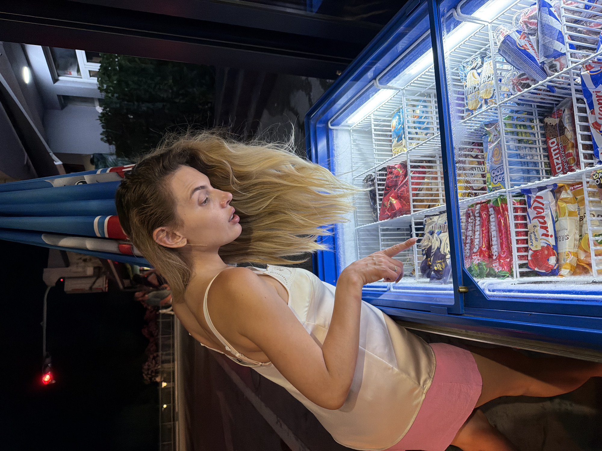 A blonde woman in a white satin camisole and a pink skirt stands in front of an open, illuminated freezer chest filled with various ice cream novelties. She is leaning forward, looking down at the selection, and pointing at one of the items with her right index finger. The freezer is inside what appears to be a small shop or convenience store, with a blue awning visible above the freezer. The background shows a street scene at night, with a traffic light displaying red and some blurred figures of people in the distance. The scene is set in Budva, Montenegro.