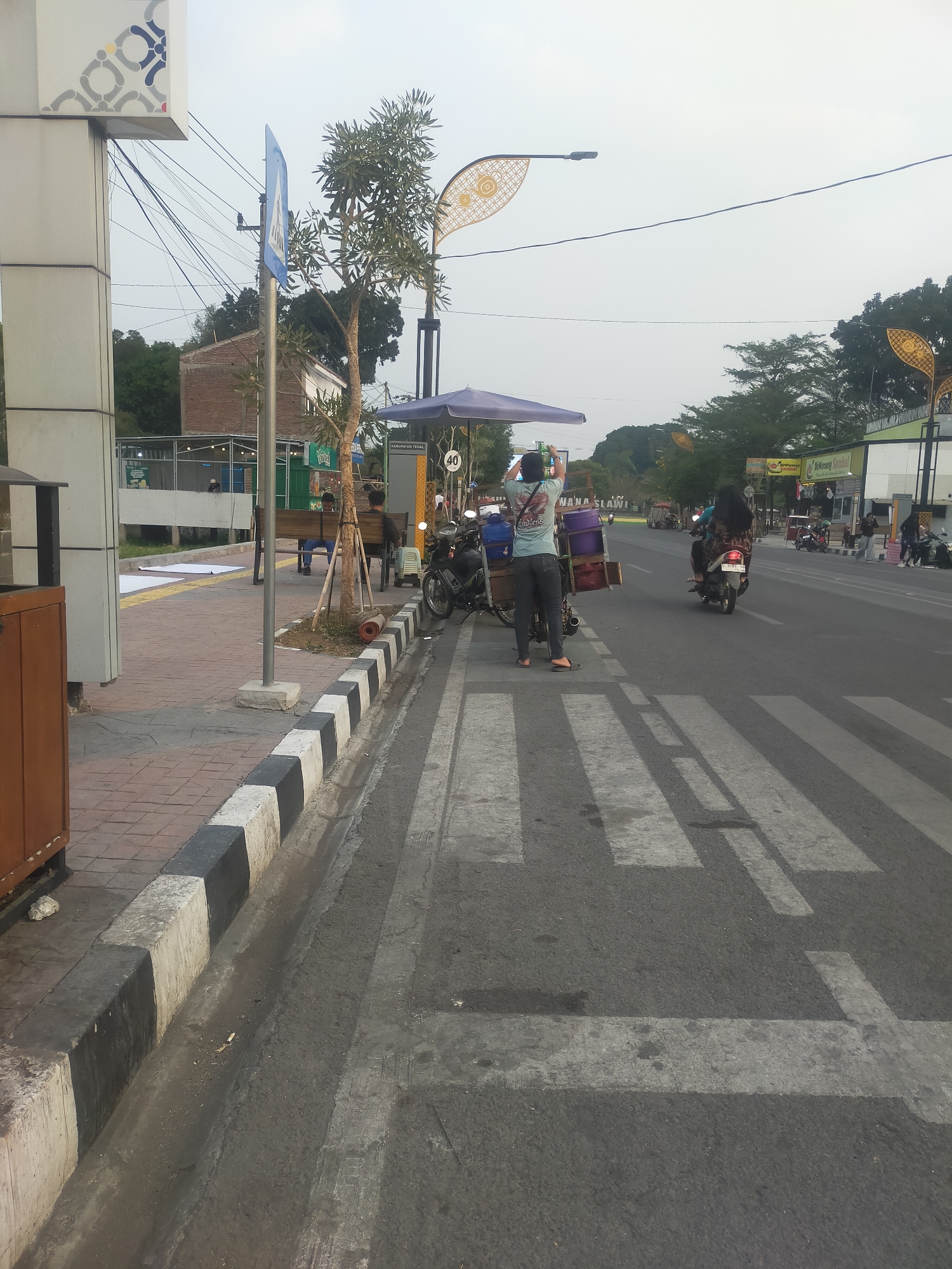 In Slawi, Indonesia, a street scene unfolds with a pedestrian crossing prominently in the foreground, marked by white stripes on asphalt. To the left, a sidewalk features a black and white striped curb. A vendor with a cart is positioned near the sidewalk, facing the street, with a motorcycle and umbrella. Behind the vendor, a few people sit on a bench. Across the street, several motorcycles are parked. Buildings line the far side of the street, and trees are visible in the background, with power lines overhead. The sky is overcast.