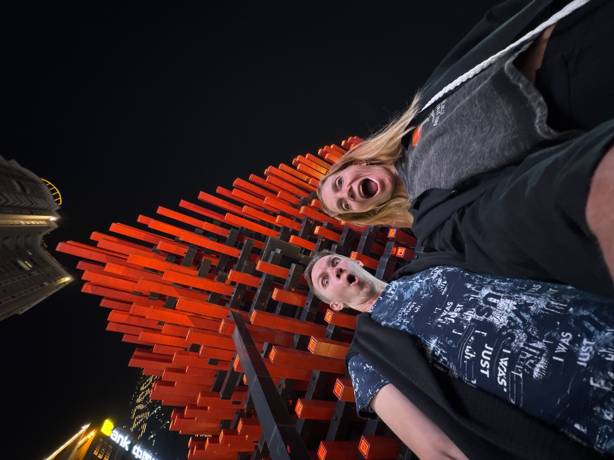 At night in Yuzhong District, China, two people, a fair-skinned woman with blonde hair and a fair-skinned man, are in the foreground looking up with exaggerated facial expressions in front of Hongyadong. The woman is screaming and the man has his mouth open in surprise. Behind them is a large, illuminated red structure. The words "Bank 中国" are visible on a nearby building. The man's shirt has the words "JUST" and "I WAS" printed on it. The buildings and structures are lit with artificial light.