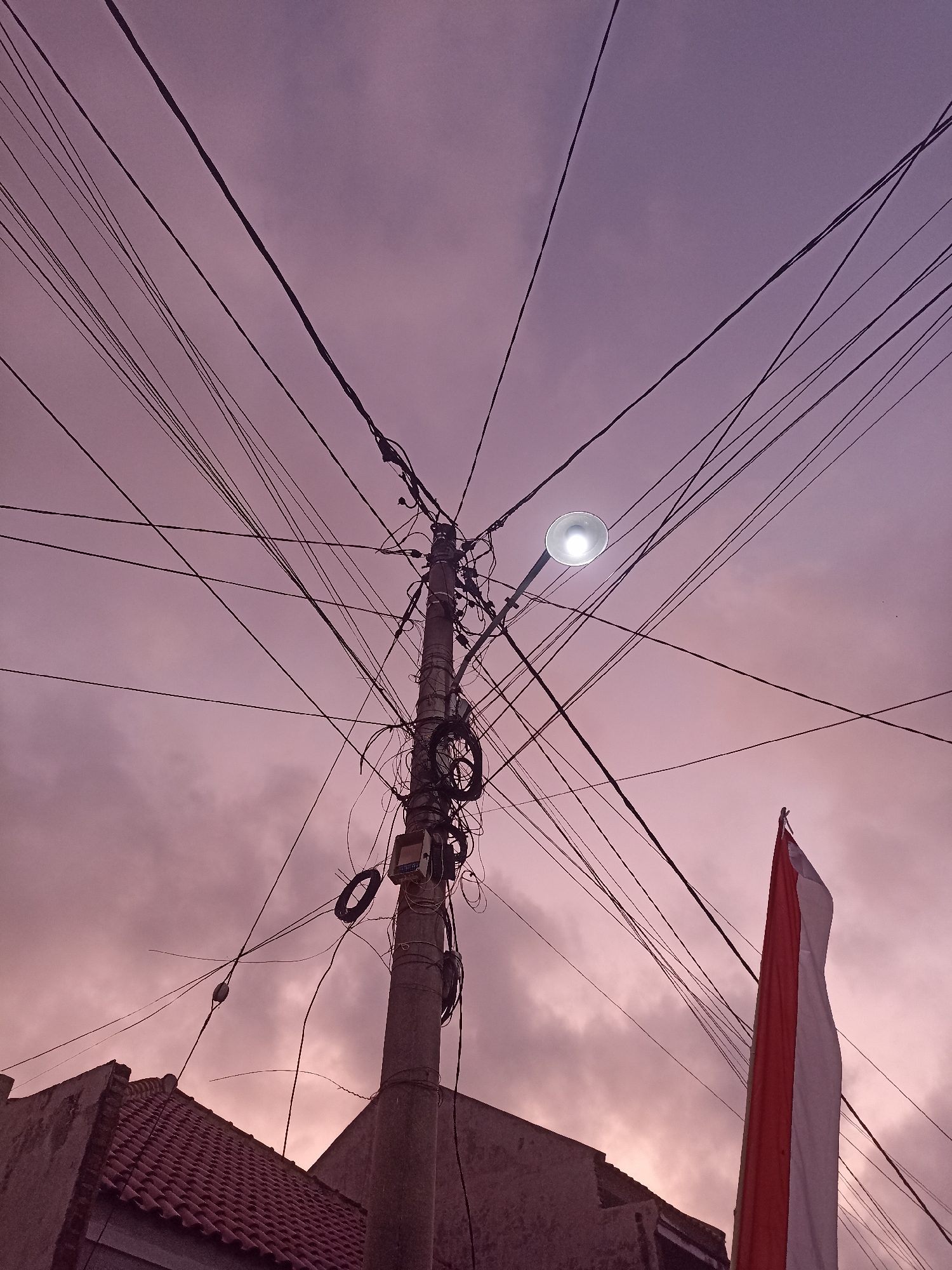 This is a low-angle, full-frame photograph of a utility pole and an Indonesian flag against a twilight sky. The utility pole, located in Mojopanggung, Indonesia, is heavily laden with a dense network of black electrical wires that crisscross the frame in various directions. A single street lamp on the pole is illuminated, casting a bright white light. Below the lamp, various electrical boxes and coiled wires are attached to the pole. To the right of the pole, a large Indonesian flag, featuring red and white horizontal stripes, is unfurled and partially obscures the sky. In the lower left corner, the tiled roof and upper portion of a building are visible. The sky is a soft gradient of purple and pink, indicating dusk or dawn, with visible cloud cover.