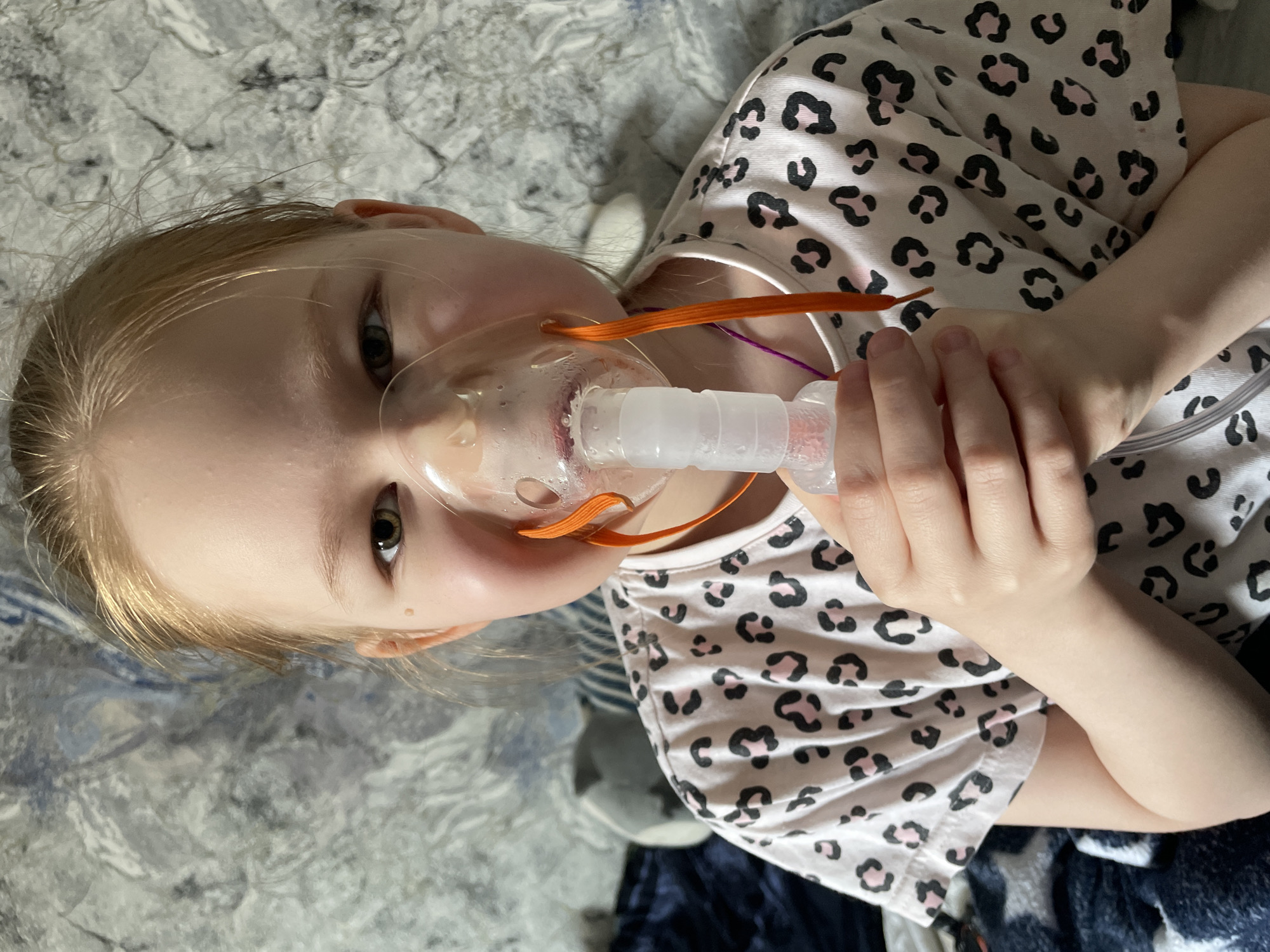 A young child with light blonde hair is shown from a top-down, slightly angled perspective, appearing to lie down in an indoor setting, likely on a bed or couch with a grey and blue patterned blanket. She is wearing a light-colored t-shirt adorned with a black and pink leopard print pattern.
The child is actively receiving a medical treatment, holding a clear plastic nebulizer mask to her face with both hands. The mask is secured by orange elastic straps, and a clear tube connects to the nebulizer cup, which shows condensation, indicating it is in operation. Her eyes are open and directed towards the viewer, displaying a calm or neutral expression.
The setting is indoors, with no discernible cues for time of day or weather. The location context provided is Noviy, Russia. There is no visible text present in the image.