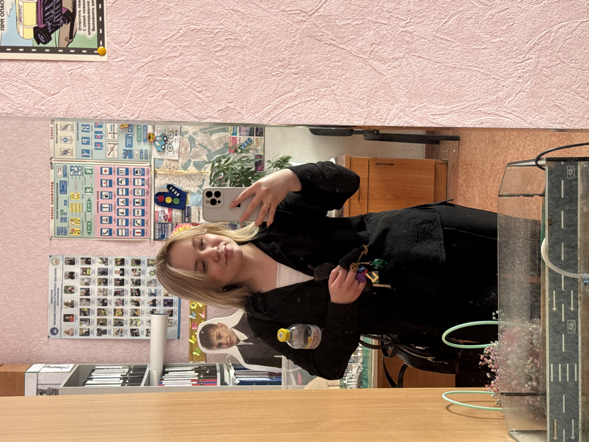 The image shows a selfie taken in a room in the city of Chaikovsky, Russia. The selfie is taken in a mirror with a light pink textured wall in the background. On the wall are traffic signs, posters, and a cutout of a person. A blond woman is holding a phone in her left hand, and some keys in her right. She is wearing a black top with a white t-shirt underneath. In the foreground, there is a wooden table. On the table, there is a magazine rack, a water bottle, a purse, and a decorative glass case with an image of a road. The mirror shows the room's contents behind her, including a wooden cabinet and a plant.