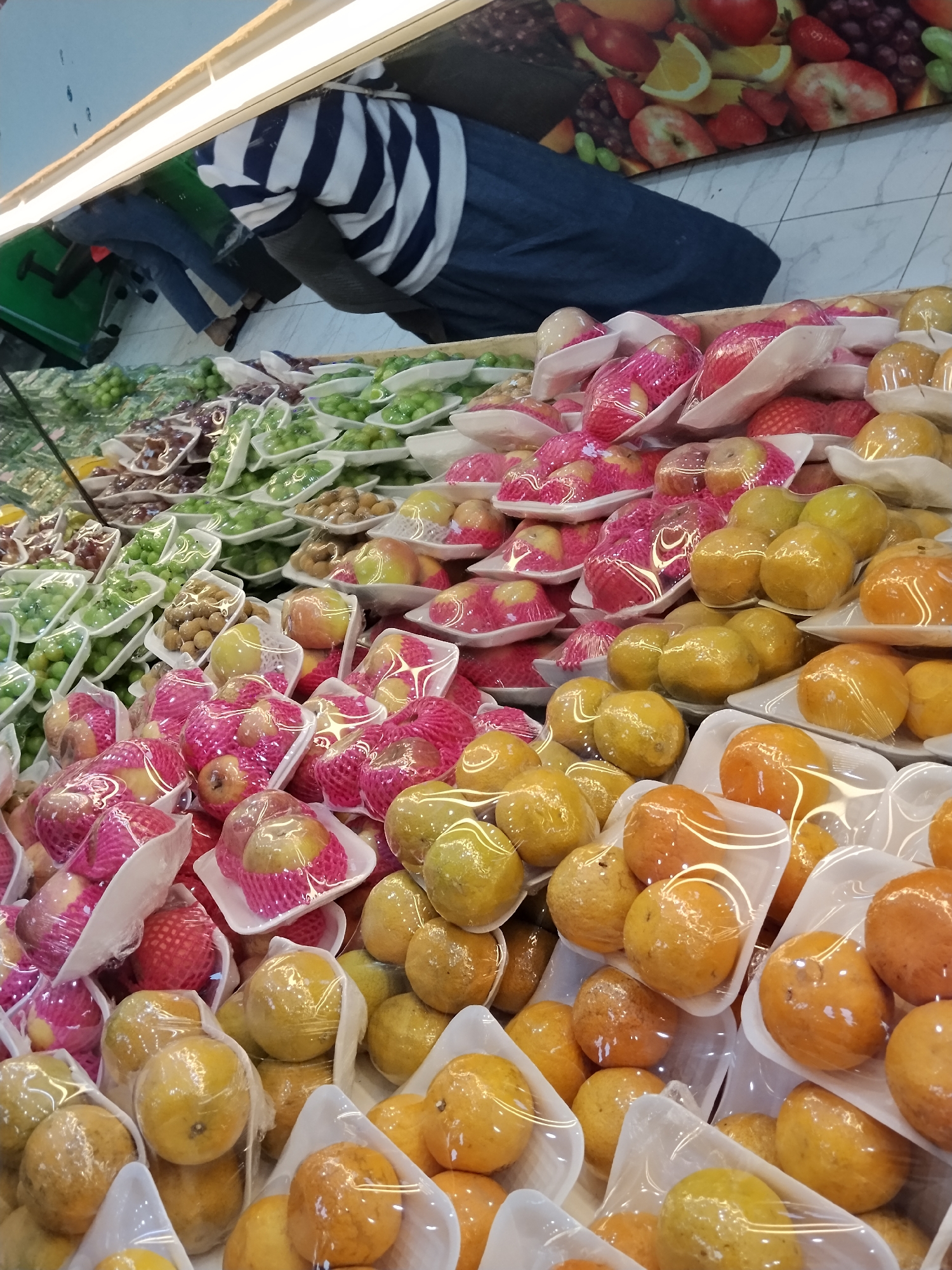 A high-angle shot captures a produce display in a supermarket, likely in Ciruas, Indonesia. The foreground is dominated by trays of various fruits, predominantly oranges and apples, all individually wrapped in plastic and housed in white styrofoam containers. Some fruits are encased in pink netting. In the background, more trays of green grapes and some darker colored grapes are visible. A person wearing a blue and white striped shirt and dark pants is partially visible in the upper left quadrant of the image, bending down, possibly attending to the produce. The lighting suggests a well-lit indoor environment.