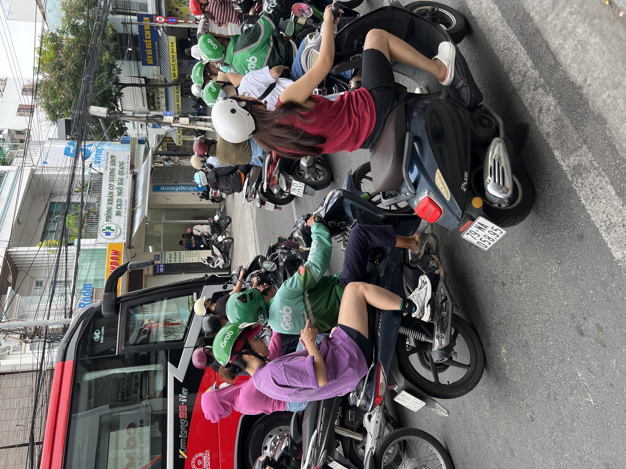 This is a street-level, eye-level shot of a busy intersection in Nha Trang, Vietnam. The image captures a moment in time where traffic is at a standstill, with numerous motorbikes and a large bus creating a congested scene. The foreground is dominated by motorbikes and their riders, many wearing helmets. A woman in a purple t-shirt and black shorts is looking down at her phone while sitting on a motorbike. Beside her, a Grab driver wearing a green uniform and helmet is also on a motorbike. In the background, a red and black bus with "Kim Long" branding is visible, along with various buildings lining the street, suggesting an urban commercial area. The overall atmosphere is one of typical city traffic in Vietnam, with a focus on motorbikes as the primary mode of transportation.
