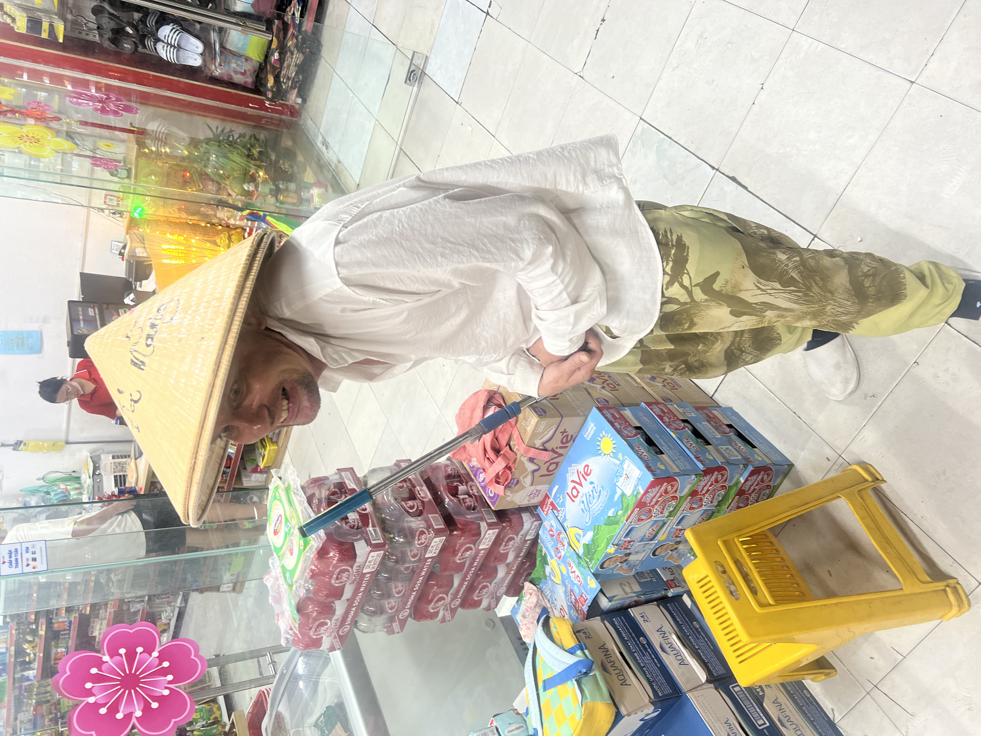 A man wearing a conical hat, a white shirt, and camouflage pants stands in a convenience store. He is smiling and holding a mop. In the background, shelves are stocked with various products, including bottled water and snacks. A cashier is visible behind a counter. The store appears to be decorated for a holiday, with pink and yellow floral accents. The man is standing next to a stack of water bottles and a yellow plastic stool. The floor is tiled in a white and grey pattern. The lighting is bright and even, suggesting an indoor setting. The image is taken from a slightly low angle, looking up at the man. The overall impression is one of a casual, everyday scene within a commercial environment.