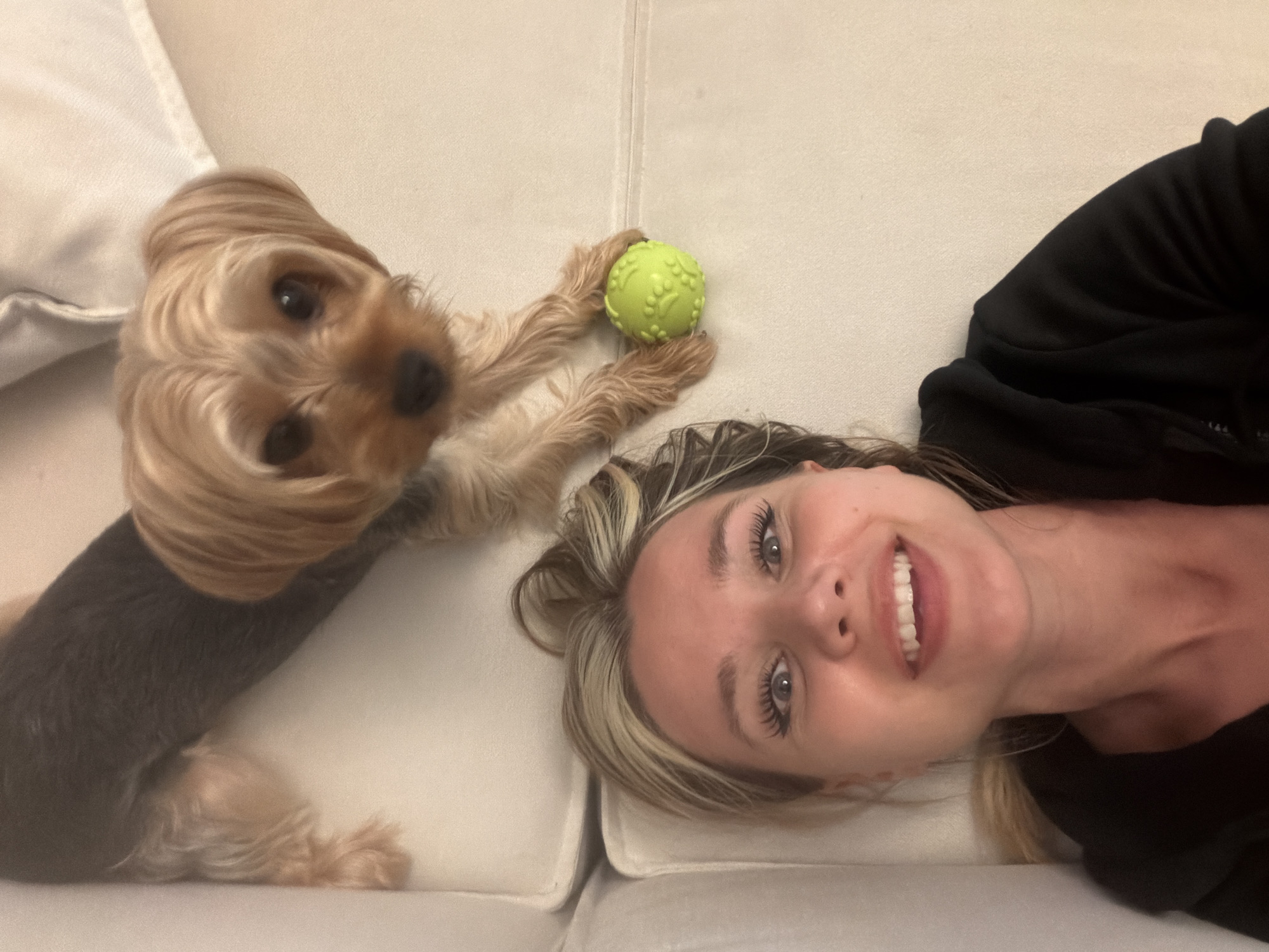 A young woman and a small, tan Yorkshire Terrier are lying on a cream-colored sofa, likely indoors. The woman is lying on her right side, with her head resting on a cushion. She has blonde-streaked brown hair, prominent eyelashes, and is smiling broadly at the camera, her mouth open to reveal white teeth. She is wearing a black hoodie. The dog is positioned to her left, also on the sofa. Its head is tilted, and its dark eyes are looking towards the camera. The dog's front paws are holding a small, bright green ball with a textured surface. The dog's fur is a mix of tan and dark brown. The scene appears to be casual and intimate, suggesting a relaxed moment between the woman and her pet. The lighting is soft and even, typical of indoor lighting, and there is no indication of the time of day or weather conditions. The setting is simple, with the focus being on the subjects and their interaction. There is no visible text in the image.