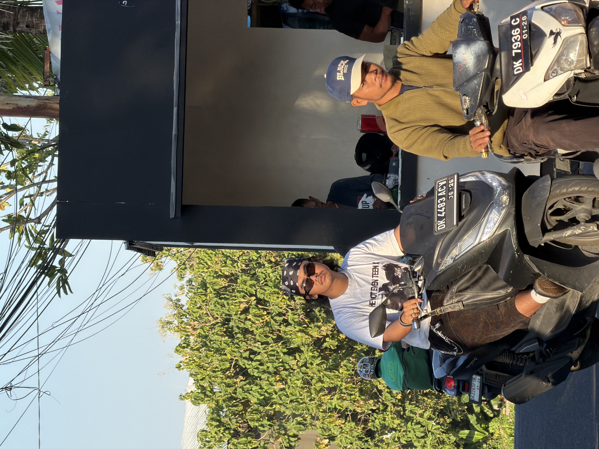 This image depicts a street scene in North Kuta, Indonesia, featuring two individuals on scooters in the foreground and a small roadside establishment in the background.
The central figure, a male, is seated on a black Honda PCX scooter, facing forward but looking slightly towards the viewer. He wears a black bandana with white paisley patterns, dark narrow sunglasses, a white short-sleeve t-shirt featuring the text "HE NOT EVEN TEEN" above a black-and-white graphic, dark brown textured shorts or pants with a metal chain accessory, white socks, and brown low-top shoes. His scooter's license plate reads "DK 4483 ACY" with "06-26" below.
To his right, another male is seated on a white scooter with visible wear on the handlebars. He wears a navy blue baseball cap with "BLACK" in white text, an olive green ribbed zip-up jacket, and dark brown pants. His scooter's license plate reads "DK 7936 C" with "01-28" below. He looks slightly to his left.
In the background on the left, behind the main figure, a person wearing a green jacket and a patterned helmet is partially visible on another scooter with license plate "DK 2265FOX" and "07-29." Behind this scooter is a dense cluster of bright green foliage.
On the right side of the image, behind the second scooter rider, is a dark gray/black, open-front structure appearing to be a small shop or stall. Inside, a light gray wall is visible, along with multiple individuals, including one male in a blue t-shirt with "UP" visible on it. Helmets and a red container are also visible inside the stall.
The sky above is a clear, light blue, crisscrossed by numerous black electrical wires. The scene is illuminated by strong sunlight, indicating either morning or late afternoon.