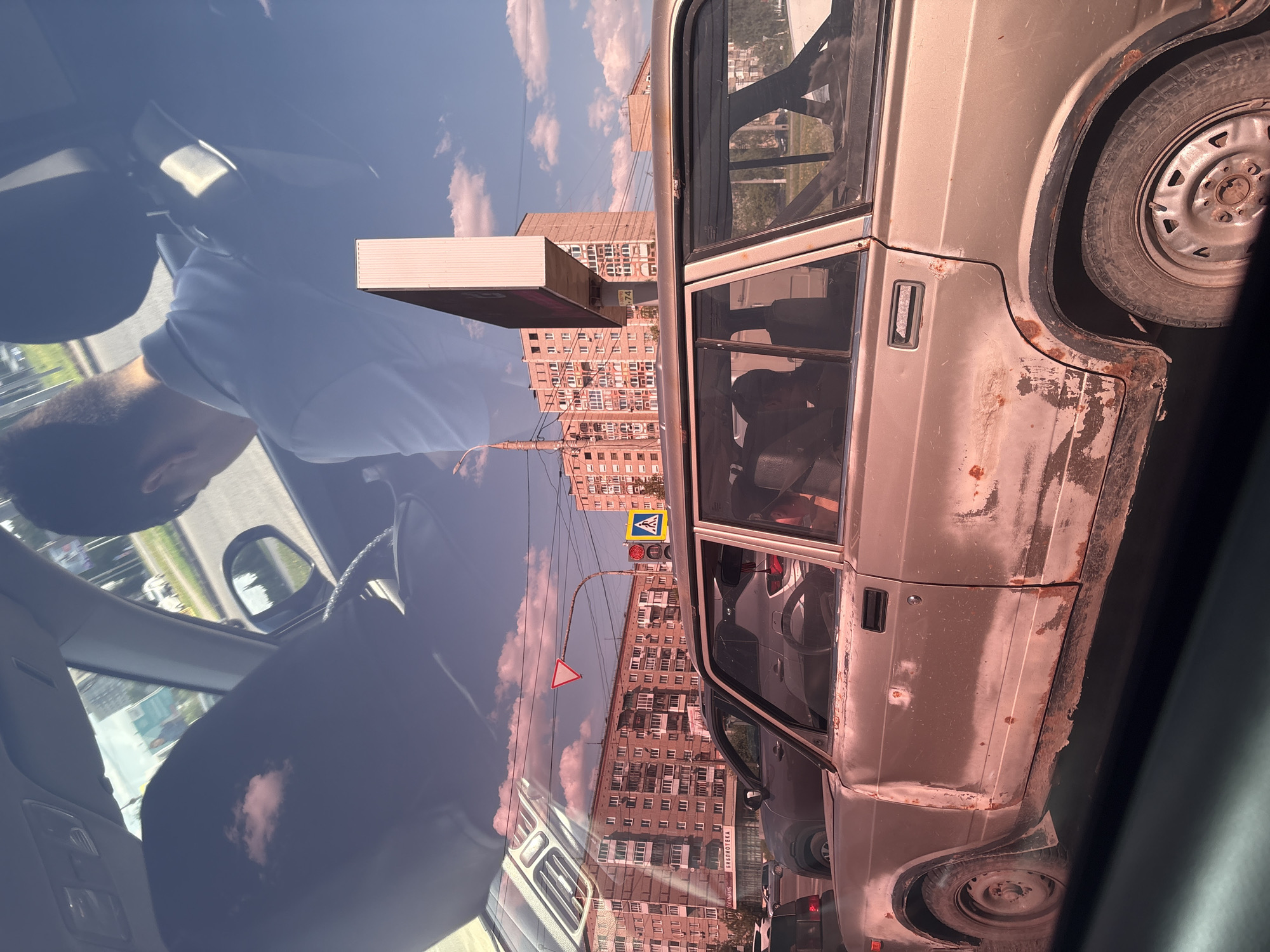 The image, taken from inside a car, shows another car adjacent in traffic in what appears to be Izhevsk, Russia. Reflected in the car's windshield is a partial view of the person taking the picture. The external car is silver and shows signs of corrosion. Pedestrian crossing signs and a building labeled "Библиотека" (Library) in Russian are visible, further suggesting a cityscape in Russia.