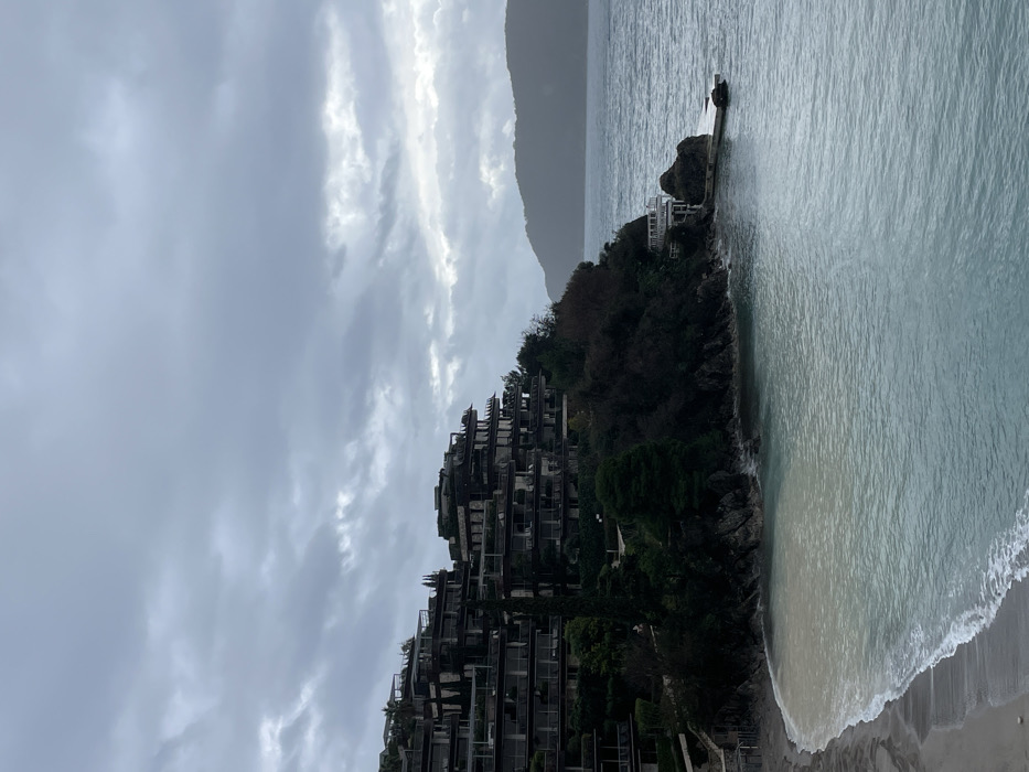 The image depicts a coastal scene on an overcast day at Sveti Stefan, in the vicinity of Budva, Montenegro. A complex of multi-story, terraced buildings with numerous balconies occupies the central and left portions of the frame, built into a steep, densely vegetated hillside. These structures are dark in tone, featuring flat roofs and an architectural style that integrates with the natural contours of the land.
In the lower-left quadrant, a light-colored sandy beach forms a crescent where small waves are breaking. The sea, extending across the lower and right sections of the image, displays a blue-green hue with some lighter, turbid areas near the shore. Along the rocky base of the cliff on the right, a small white-railed structure, possibly a deck or pavilion, connects to a narrow concrete pier or jetty that extends into the water, adjacent to a prominent dark rock formation. Distant, dark hills or mountains are visible on the far-right horizon, partially obscured by atmospheric haze or precipitation, blending with the uniformly grey, overcast sky. No individuals or active interactions are visible within the scene.