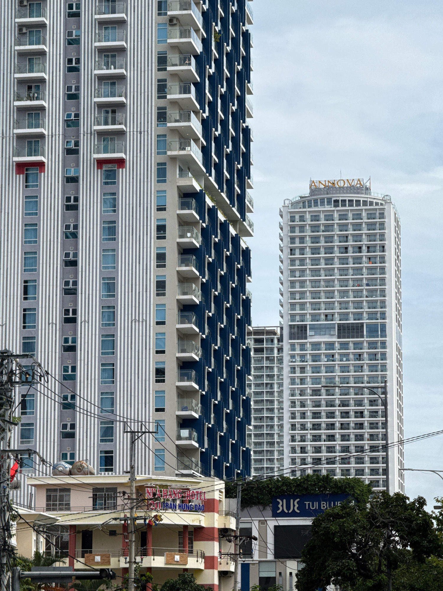 In this medium shot captured on a cloudy day in Nha Trang, Vietnam, three high-rise buildings dominate the frame. The tallest building on the right, a modern white structure, features a curved facade and is topped with an orange sign that reads "ANNOVA." This building is densely packed with balconies and windows, suggesting it is a hotel or residential complex. To its left, a second high-rise building is visible, characterized by dark blue panels and numerous balconies. The leftmost building is a tall, rectangular structure with a light grey facade accented by vertical white stripes. This building also has many balconies and windows. In the foreground, a lower building with a pink, yellow, and red striped facade is partially obscured by a dense network of electrical wires and utility poles. A sign on this lower building reads "NHÀ HÀNG HOTEL 221 TRẦN HƯNG ĐẠO." Further to the right and lower down, another building displays a blue sign with white lettering that reads "BLUE TUI BLUE." Green trees and foliage are present in the lower right portion of the image.