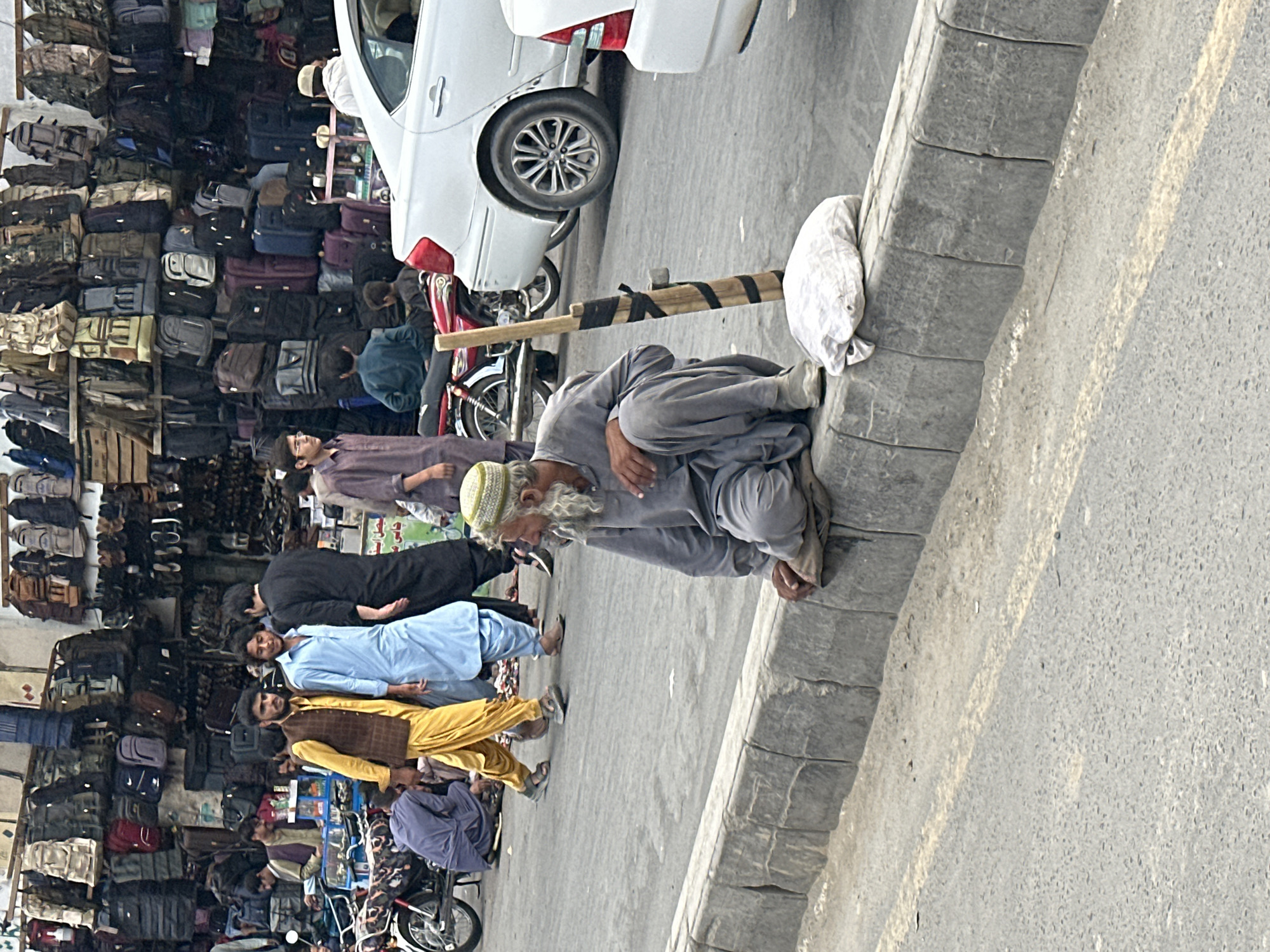 The image captures a street scene in Quetta City Tehsil, Pakistan. A man wearing a gray outfit is kneeling on the sidewalk. Behind him, other men are standing and sitting near a shop. A white car is parked beside the kneeling man. Numerous bags are displayed on the wall of the shop. A motorcycle is also present in the scene. The environment appears to be a bustling commercial area.