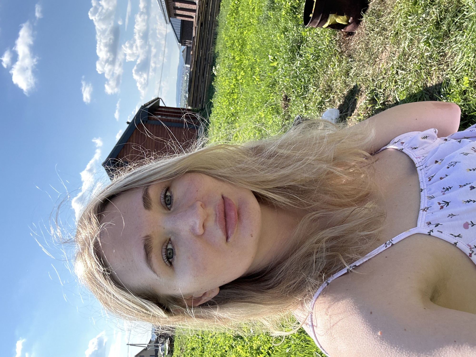 A young Caucasian woman with blonde hair and fair skin is taking a selfie outdoors on a sunny day. She is wearing a light-colored dress with a floral pattern. Her eyes are green, and she has a mole on her cheek. The background shows a grassy hill with several wooden structures, likely houses or sheds, and a stack of lumber. The sky is blue with scattered white clouds. The location is identified as Mokino, Russia, implying a rural or semi-rural setting.