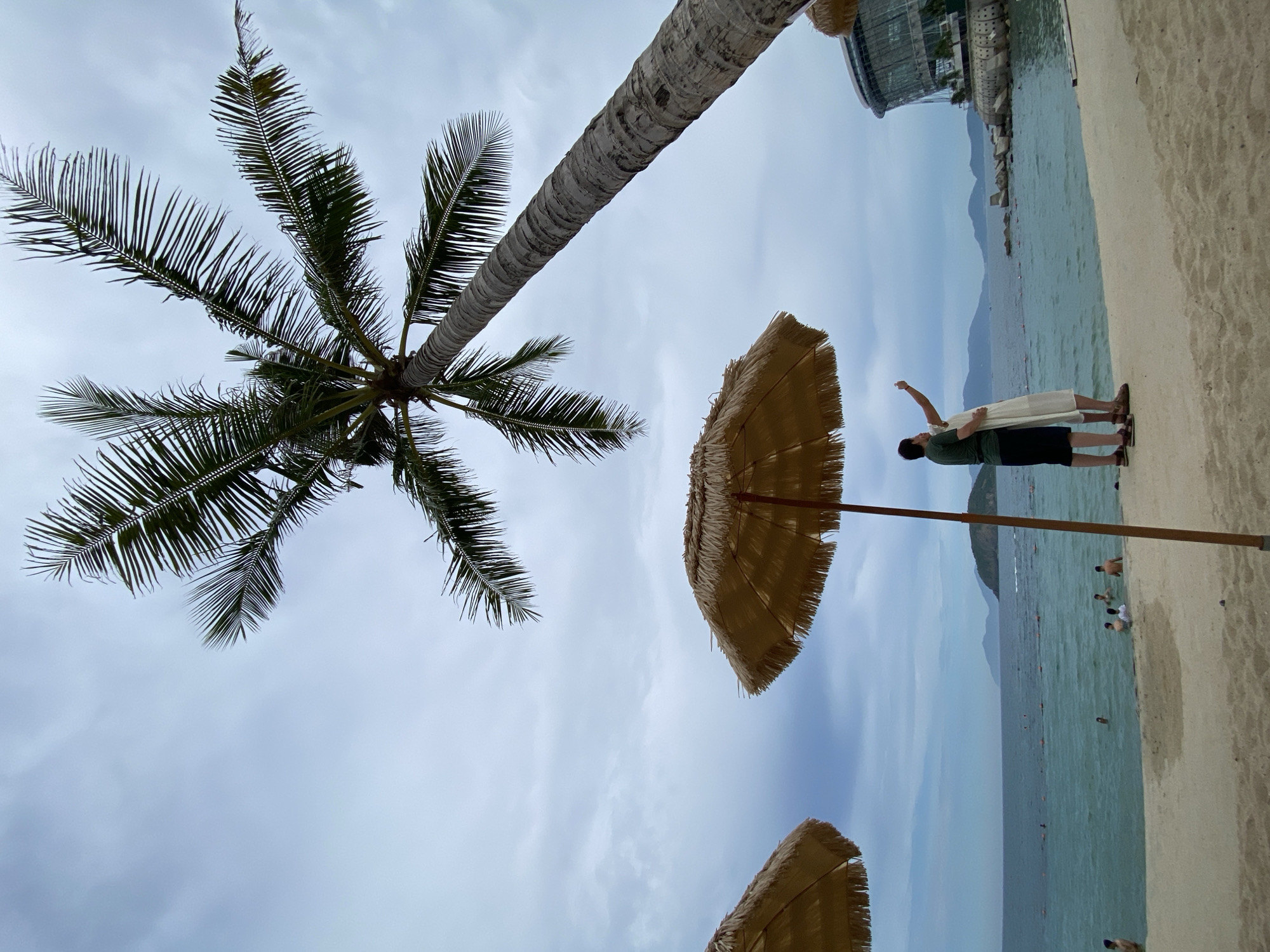 This image depicts a coastal scene in Nha Trang, Vietnam, likely near the Vinpearl Land resort area given the distinctive modern glass building visible on the right. A tall, mature palm tree dominates the upper portion of the frame, its fronds reaching across a cloudy sky. In the foreground, a sandy beach meets the calm, turquoise ocean. Two people, a man and a woman, stand on the beach, facing the water and appearing to take a selfie. The woman is wearing a white dress, and the man is in a dark green t-shirt and black shorts. Two thatched beach umbrellas are positioned on the sand, one in the left foreground and another more centrally. The ocean is populated with several individuals swimming and wading, with buoys marking a designated swimming area. Distant islands form a hazy backdrop against the horizon. The overall atmosphere is relaxed and tropical, despite the overcast sky.