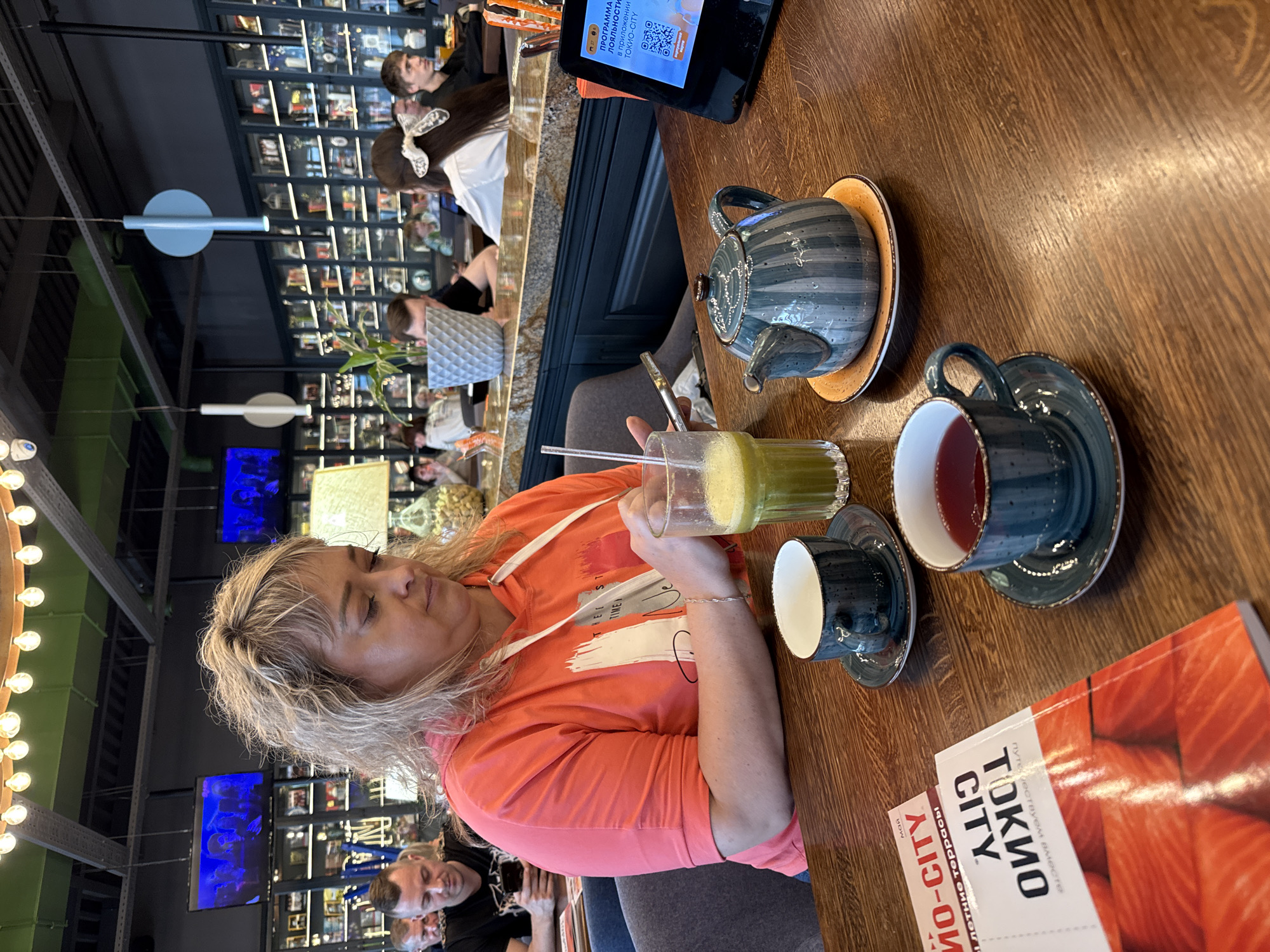 The image shows a woman sitting at a wooden table in a restaurant. She is wearing an orange hoodie and holding a glass of green juice. On the table are a teapot, two teacups, and a book titled "Tokyo City". In the background, there are other people in the restaurant and shelves filled with objects, resembling a bookstore or a cafe with a bookstore theme. The QR code on the tablet indicates the location in Saint Petersburg, Russia.