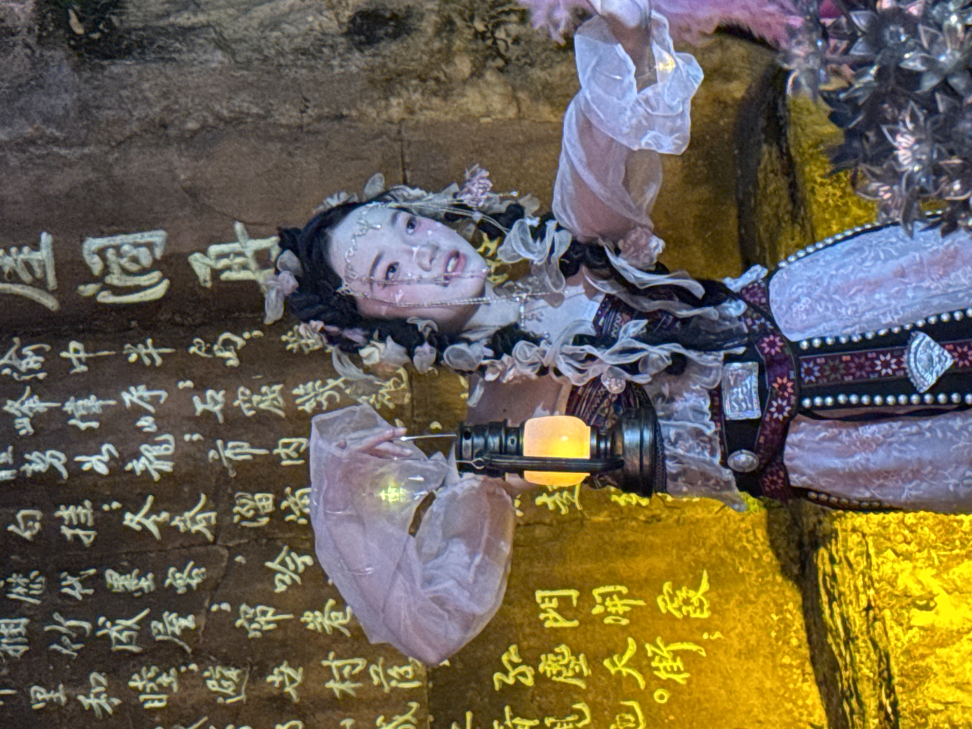 In Yuzhong District, China, a young woman with elaborate hair ornaments and face paint, dressed in traditional Chinese attire, holds an illuminated lantern. The lantern, with a dark metal frame and glowing orange light, hangs from a string. The woman's costume features a pink and white patterned dress, a dark belt with decorative buckles, and translucent sleeves. Behind her, a rough, textured stone wall is inscribed with yellow Chinese characters. The lighting is dramatic, with the wall and the woman's lower body cast in a strong yellow glow.
