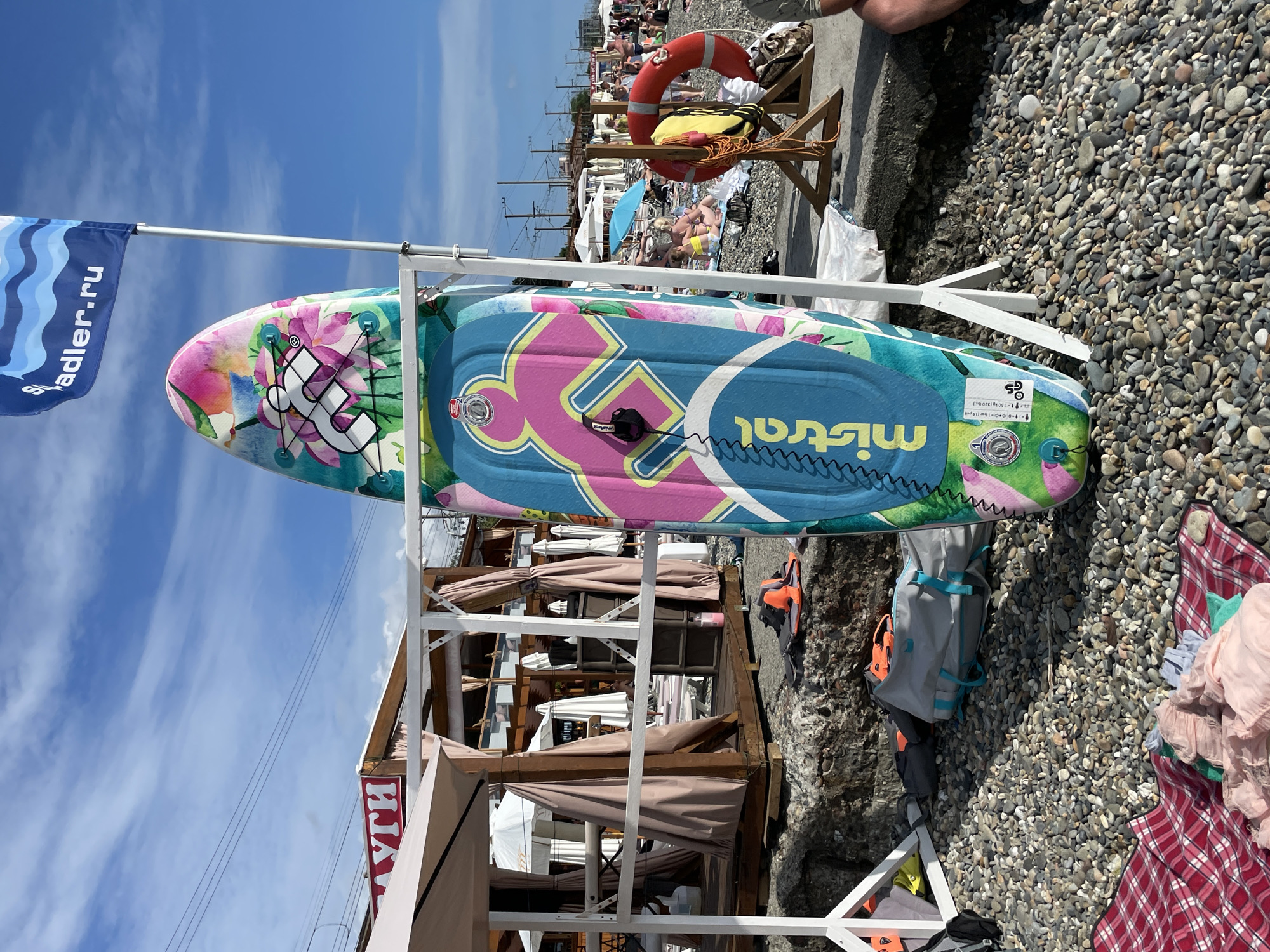 An outdoor scene on a sunny day at Adler beach in Sochi, Russia. The foreground is a pebble beach with a red and white checkered blanket and folded clothes. Two paddleboards are propped up on a white rack. The top paddleboard has a floral design with pink and green colors. The bottom paddleboard is blue with a pink and yellow graphic design and the brand name "mistral" in yellow lettering. To the right of the paddleboards, a red lifebuoy hangs on a wooden stand. In the midground, a beach cafe structure with thatched roofs and brown curtains is visible, along with beach umbrellas and loungers. Several people are sunbathing and relaxing on the beach. In the background, there are trees and buildings, and a blue sky with wispy clouds. A flag with a wave design and the text "adler.ru" is attached to a pole.