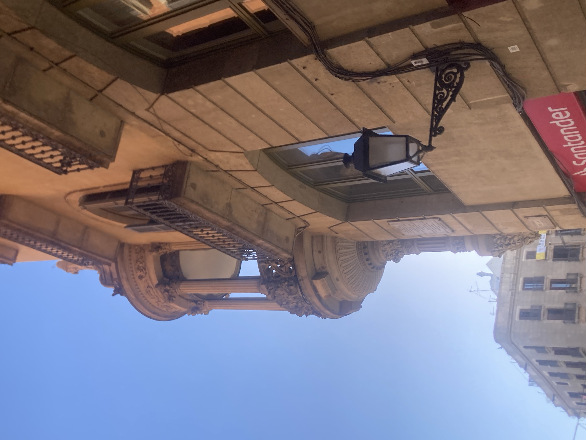 The image shows a close-up, upward-angle view of an ornate building facade against a clear blue sky. The building features a curved balcony with intricate iron railings and classical architectural elements like columns and carved details. To the right, a window with reflections of the sky is visible, and a black wrought-iron lantern is mounted on the wall, connected by a bundle of dark cables. Below the lantern, a bright pink sign with the white text "Santander" is partially visible. In the background, a lower building with multiple windows and balconies is visible, suggesting an urban setting. The overall impression is of an old European city, likely Barcelona, given the architectural style and the presence of the Santander bank.