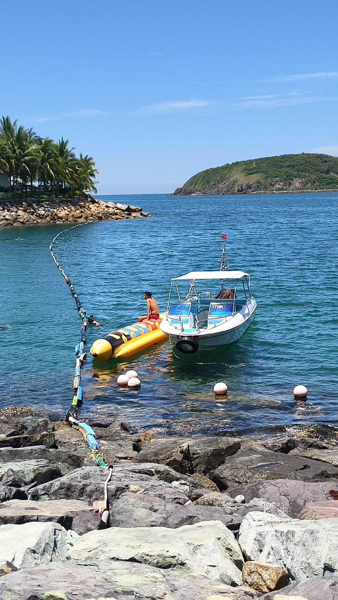 In a coastal scene at an unidentified location, a man in red shorts sits on a yellow inflatable banana boat. The banana boat is tethered to a floating line marked with multi-colored buoys. A white boat with a canopy is docked next to the banana boat, displaying "TT14K" on its side. The water is clear and blue, with a rocky shoreline in the foreground. Palm trees and a lush green island are visible in the background under a clear blue sky.