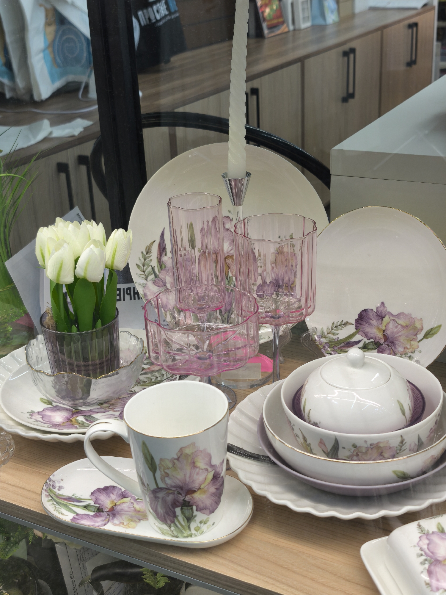 The image displays a collection of white ceramic dishes and glassware adorned with purple iris floral patterns, arranged on a wooden surface. A white candle in a clear candlestick stands prominently in the background, behind a stack of pink glass goblets. In the foreground, a white ceramic mug sits on a matching saucer, both decorated with the same iris design. To the left, a small arrangement of white artificial tulips in a grey ribbed glass vase adds a touch of greenery. The dishes include dinner plates, a lidded serving bowl, and smaller bowls. The overall scene suggests a table setting or a retail display of tableware. The reflection in the glass surface in the background shows shelves with various items, including books and what appears to be a poster with Russian text reading "ПРОСИМ" (prosim), meaning "please" or "you're welcome." The environment appears to be indoors, possibly a shop or a dining area. There are no people or animals visible in the image. The city of Yoshkar-Ola, Russia, is not visually identifiable within this image.