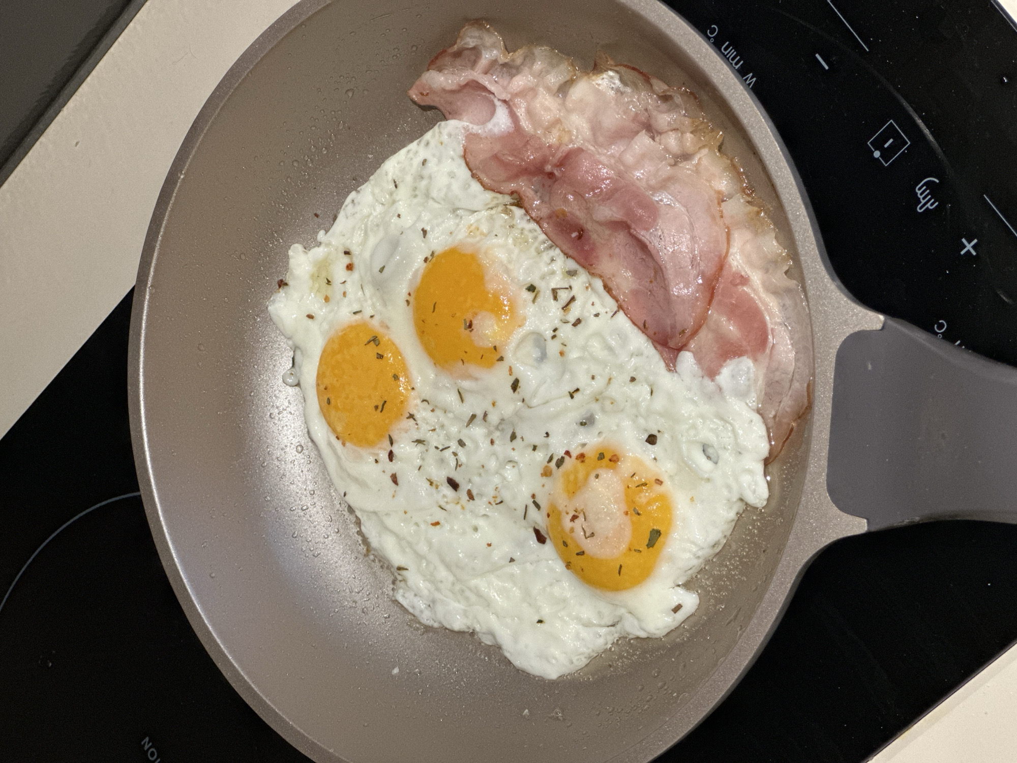 A top-down shot reveals a skillet resting on a black induction stovetop, indicating an indoor setting. Three fried eggs with bright yellow yolks and cooked egg whites are sprinkled with herbs. A few strips of uncooked bacon rest on the top edge of the eggs. The skillet has a gray exterior and a gray handle. The stovetop features touch-sensitive controls on the right side of the skillet. No people are visible in the image. The location is not discernible from the image, therefore the city of Batumi, Georgia is not determined.